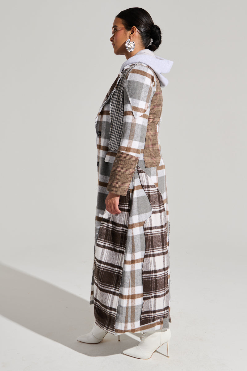 A person stands in profile wearing the MIXED PLAID NOT FEELINGS PATCHWORK TRENCH featuring tortoiseshell button closures over a white hoodie, accented with large earrings and white high-heeled ankle boots. The background is plain and neutral.