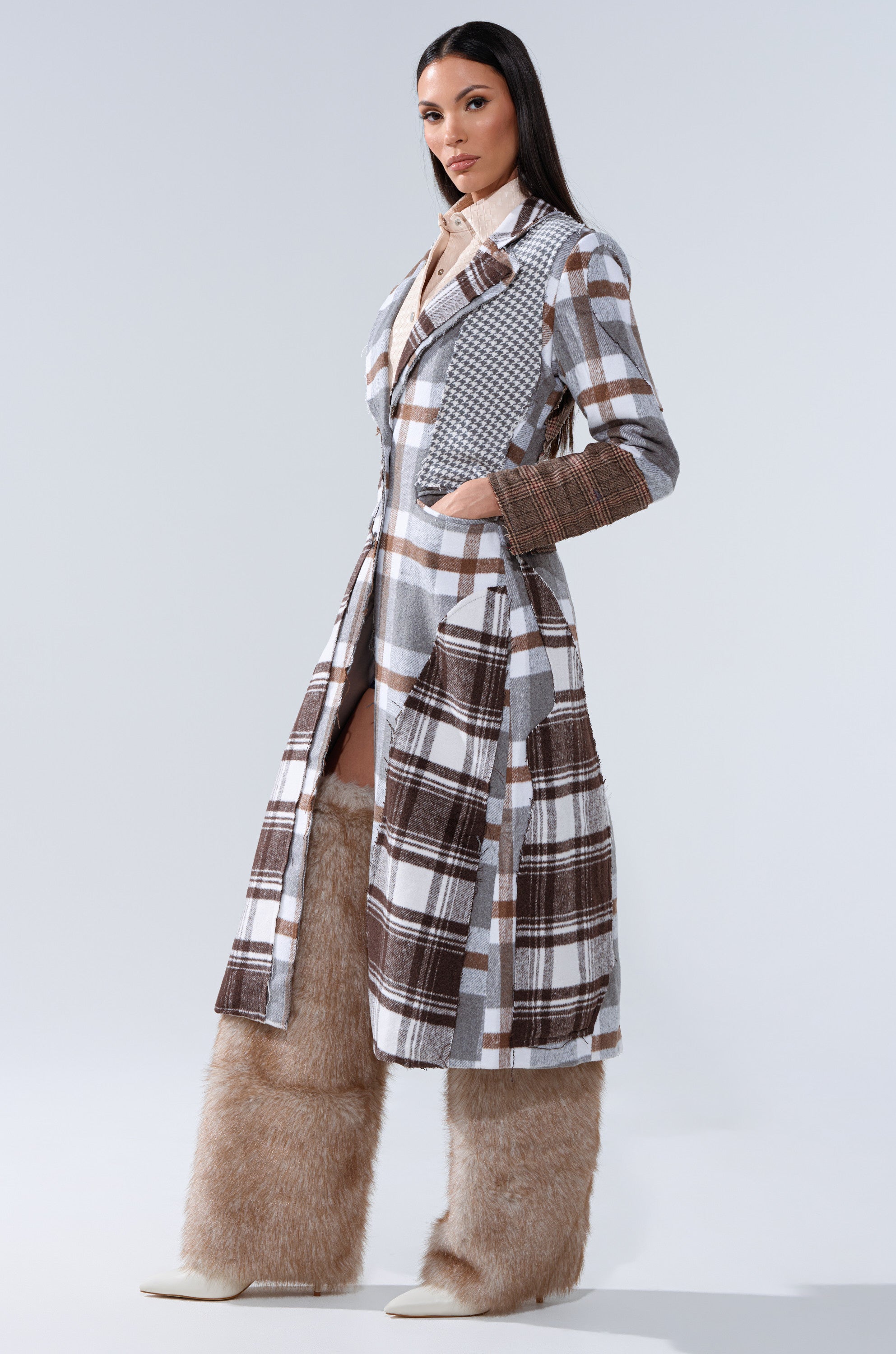 A woman wears the MIXED PLAID NOT FEELINGS PATCHWORK TRENCH, featuring gray and brown patterns, with beige furry pants and white pointed shoes against a plain light gray background.