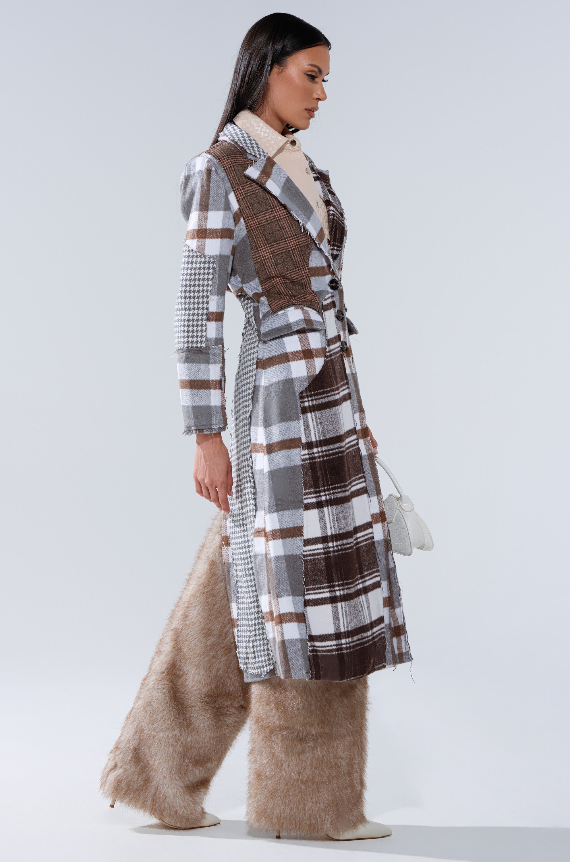 A woman in profile wears the MIXED PLAID NOT FEELINGS PATCHWORK TRENCH over wide-legged beige faux fur pants and white heeled boots, holding a small white handbag, set against a plain light background.