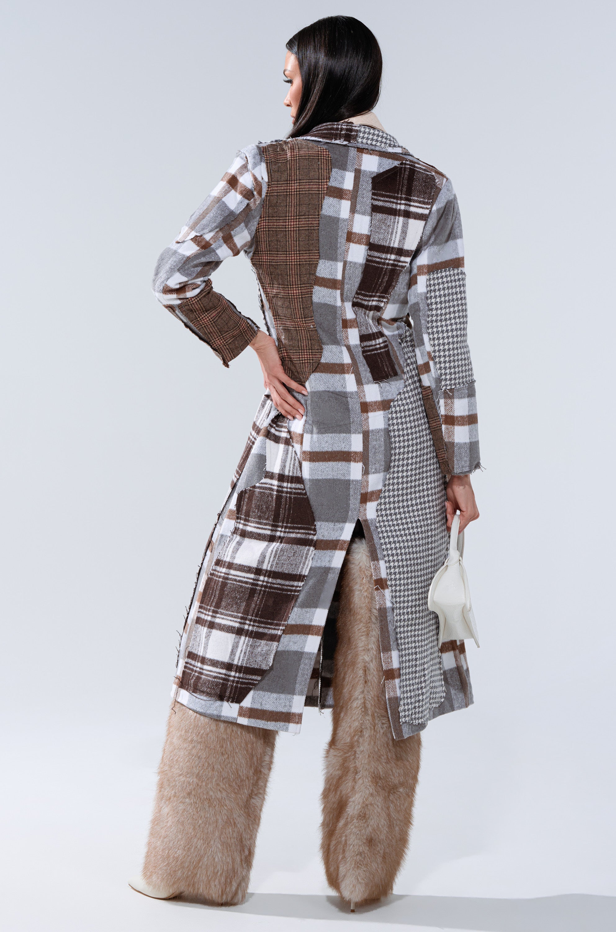 A woman, seen from the back, wears the MIXED PLAID NOT FEELINGS PATCHWORK TRENCH with brown, gray, and white plaid and houndstooth patterns, tan furry pants, and carries a small white handbag in her left hand.