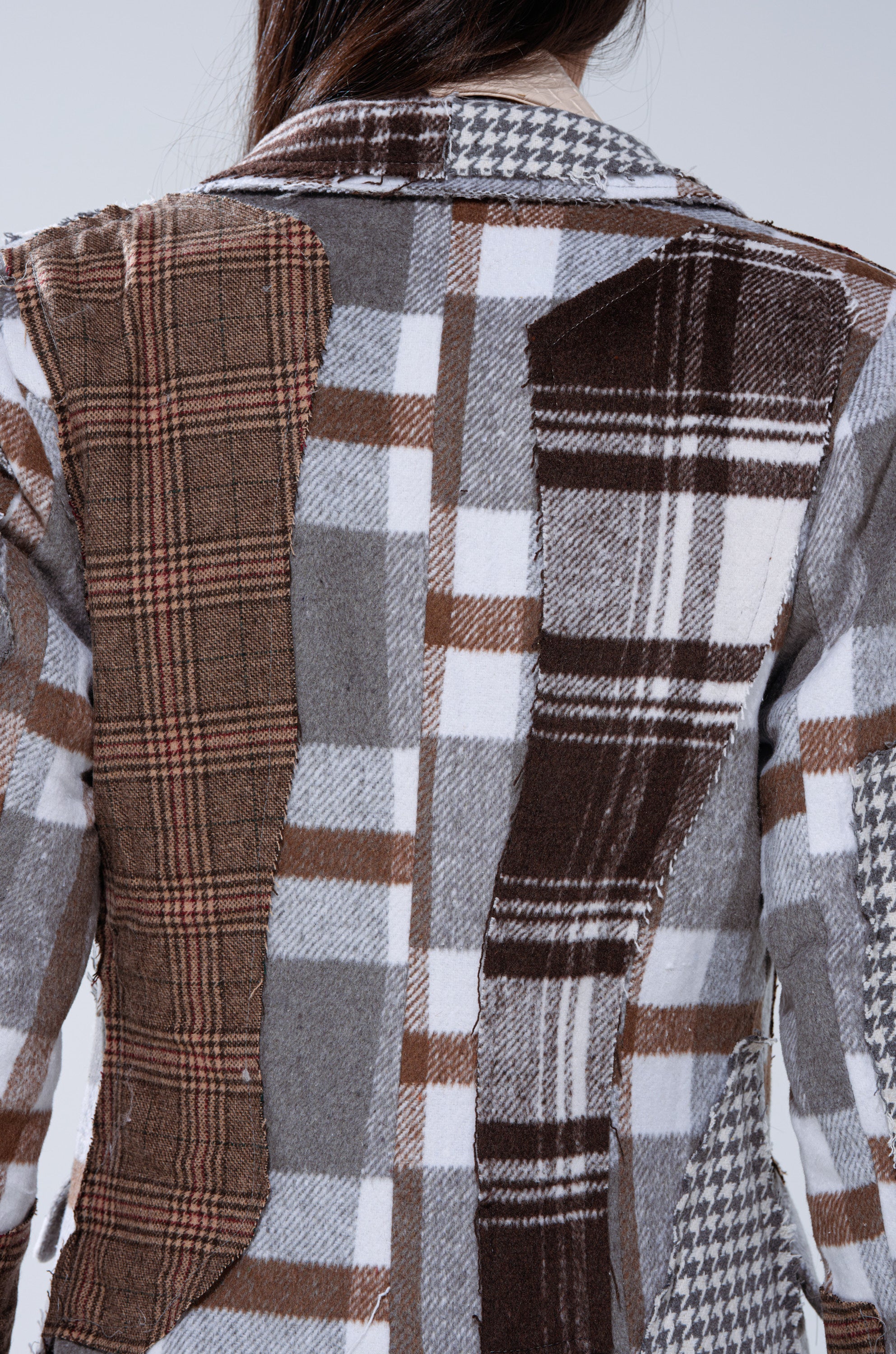 A person seen from the back wears the AKIRA LABEL MIXED PLAID NOT FEELINGS PATCHWORK TRENCH, featuring brown, gray, and white plaid and checkered fabric patches.