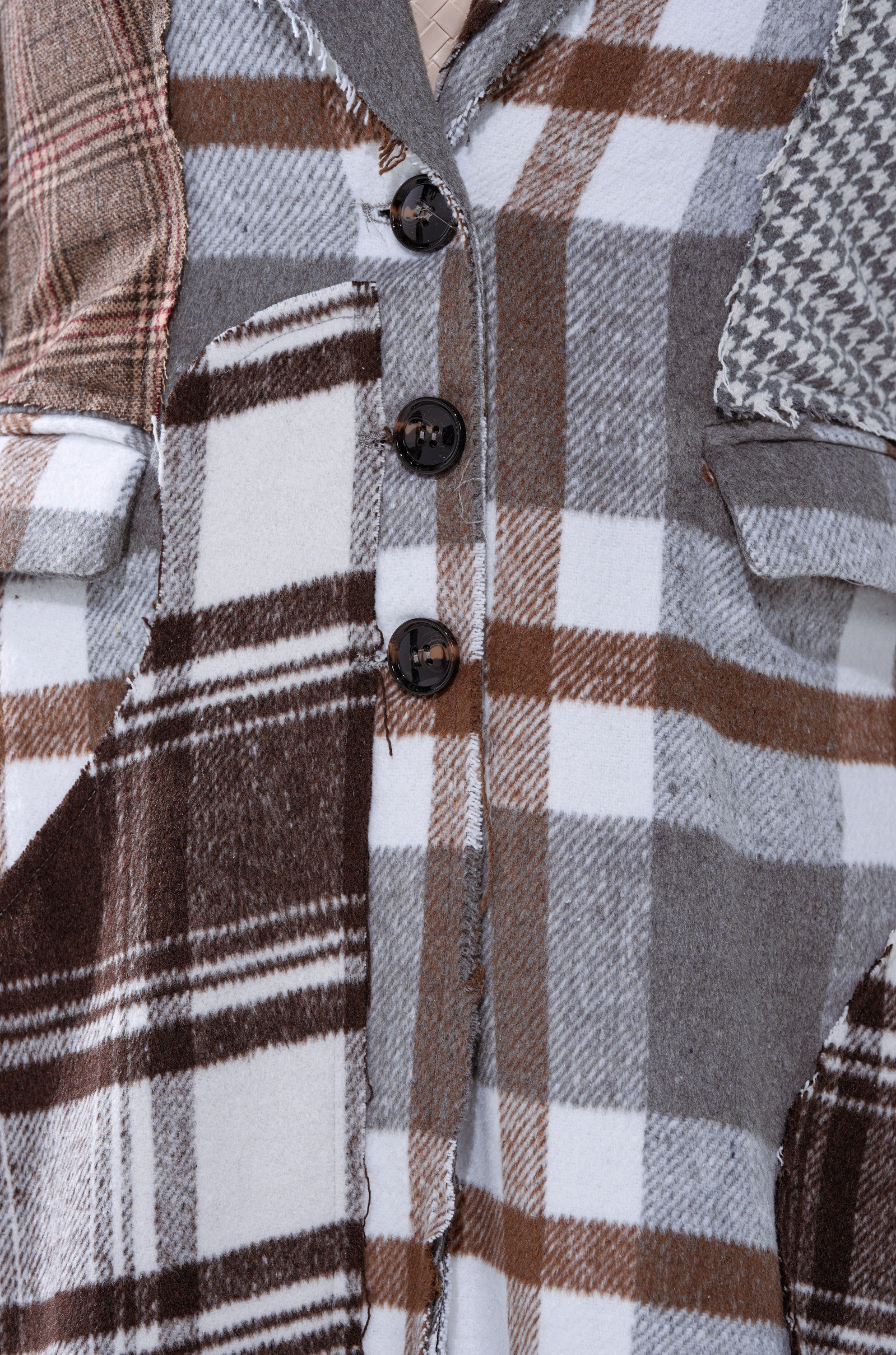 A close-up of the MIXED PLAID NOT FEELINGS PATCHWORK TRENCH, featuring plaid and houndstooth patterns in brown, gray, and white with three dark center buttons.