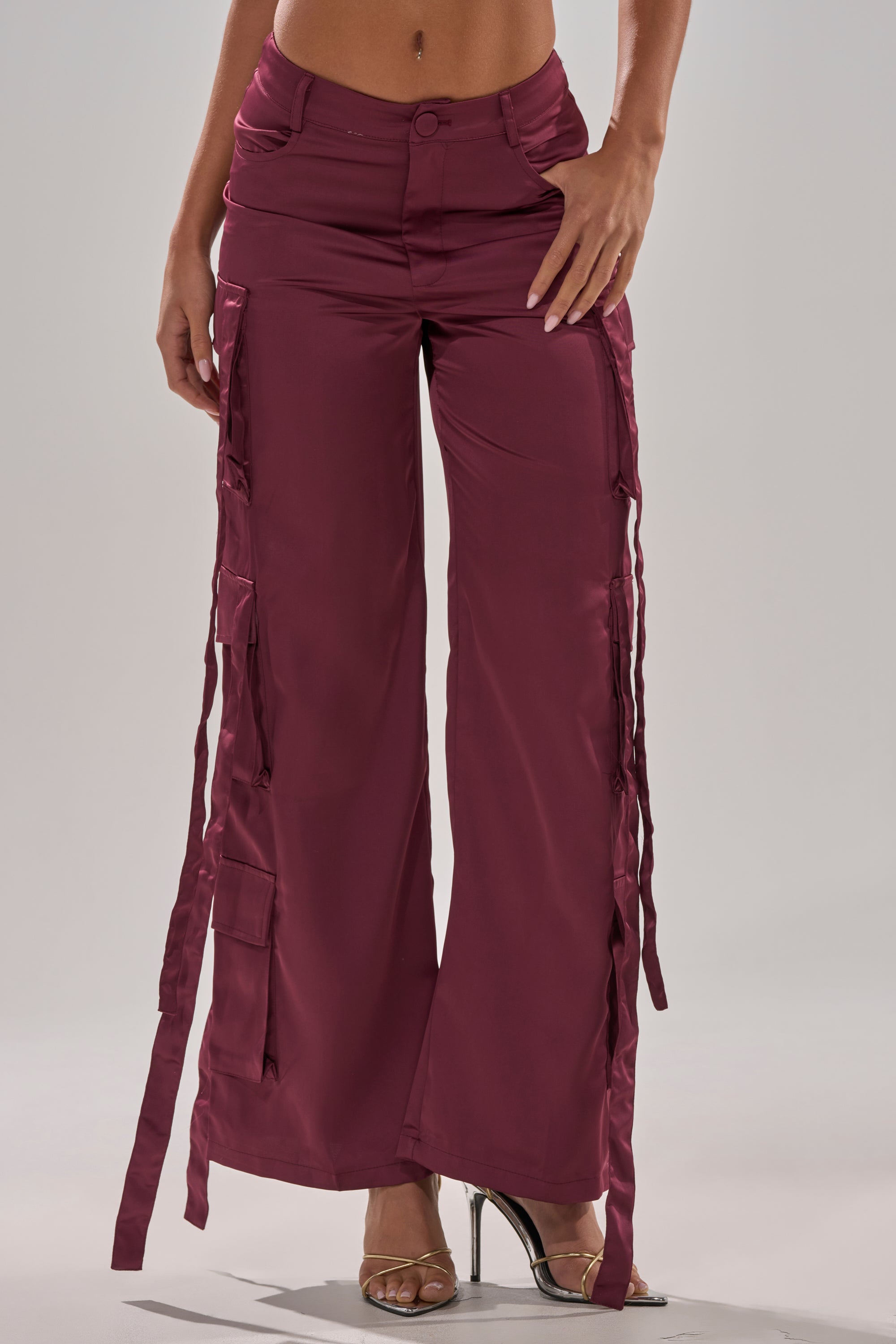 Someone wears the TAKE ME EVERYWHERE SATIN CARGO PANT in maroon, featuring a high-rise waist, wide-leg fit, side straps, and pockets, styled with clear high-heeled sandals against a plain light background. 1