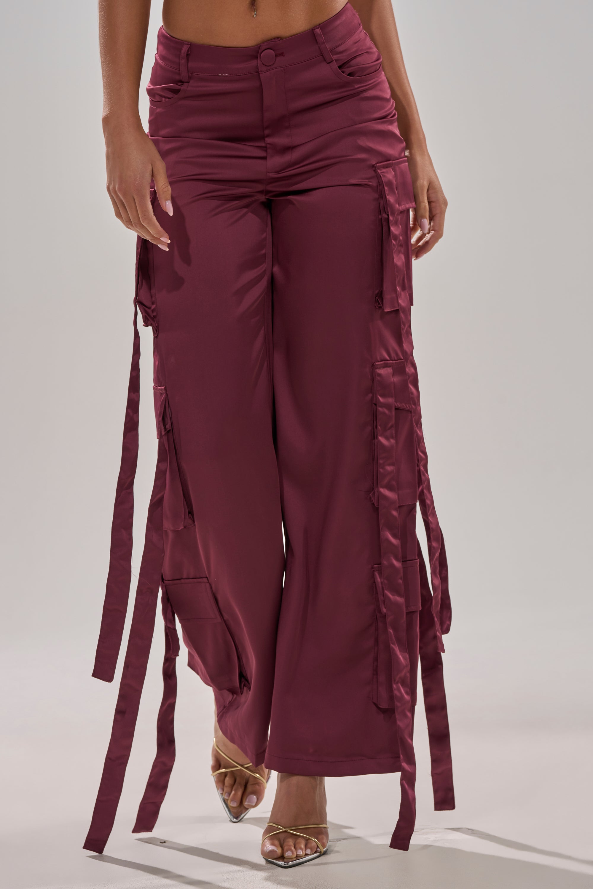 Someone wears the TAKE ME EVERYWHERE SATIN CARGO PANT, featuring a high-rise waist, wide legs, maroon satin fabric, and ribbon-like side straps, paired with clear high-heeled sandals against a plain light background. 2