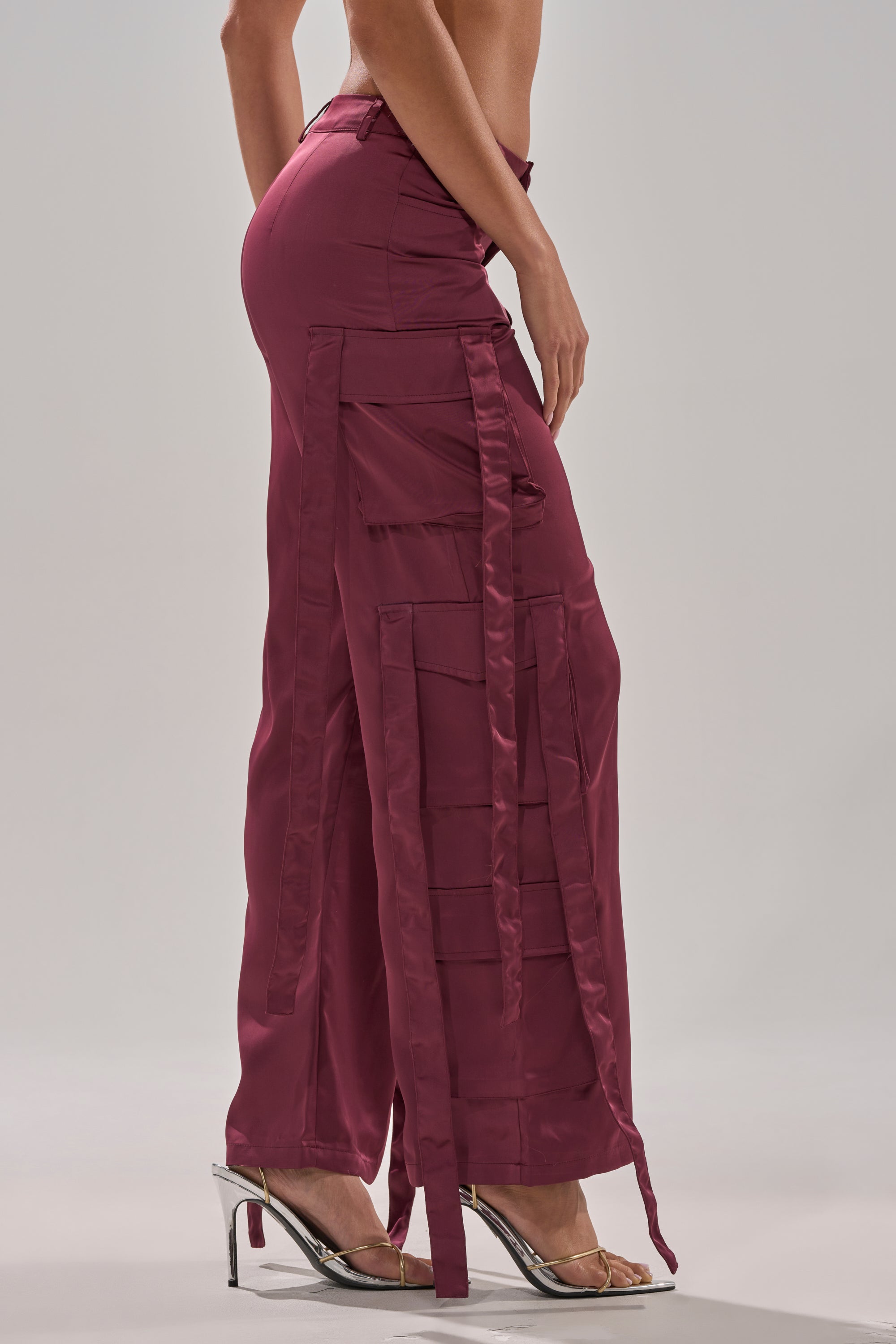 A model showcases the TAKE ME EVERYWHERE SATIN CARGO PANT in wine, featuring a high-rise waist, wide-leg silhouette, and multiple pockets, styled with clear heels against a plain light background. 3