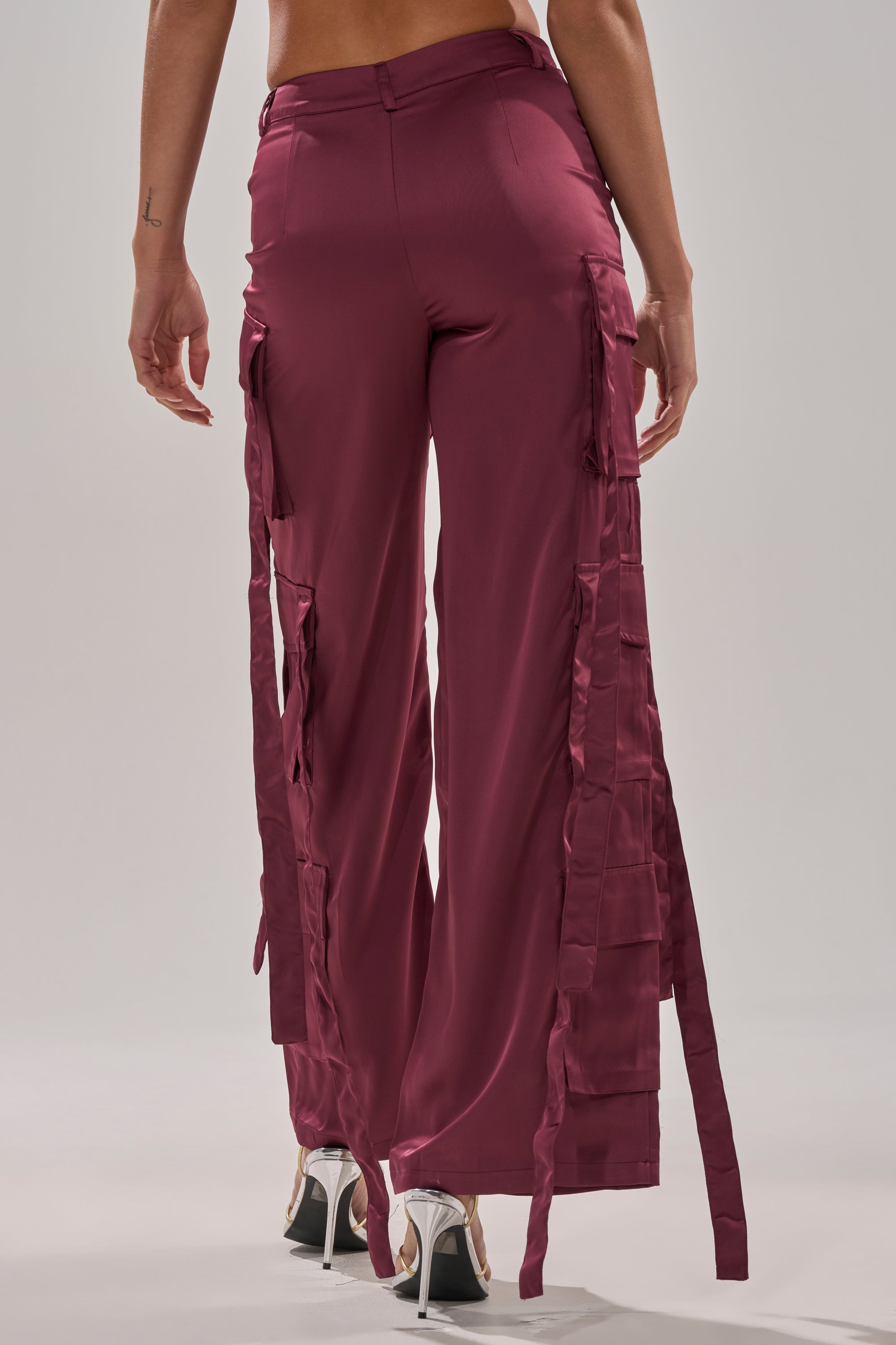 A person shown from the back wears TAKE ME EVERYWHERE SATIN CARGO PANT, featuring a high-rise wide-leg fit in mauve with ribbon details, paired with high-heeled sandals against a plain background. 4