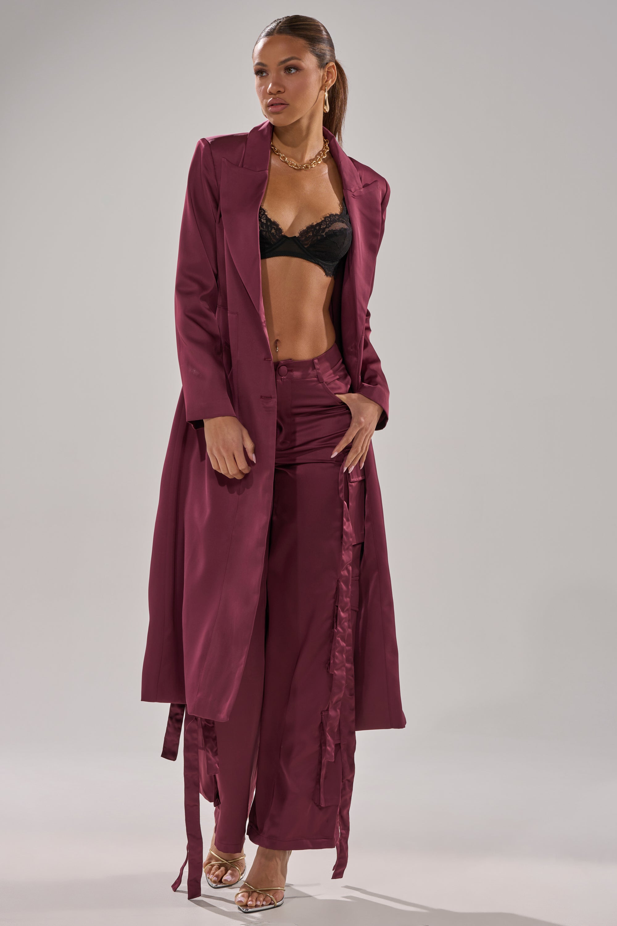 A woman poses in the TAKE ME EVERYWHERE SATIN CARGO PANT, styled with a matching long burgundy satin coat, black lace bra, gold chain necklace, and heeled sandals against a plain light gray background. 5