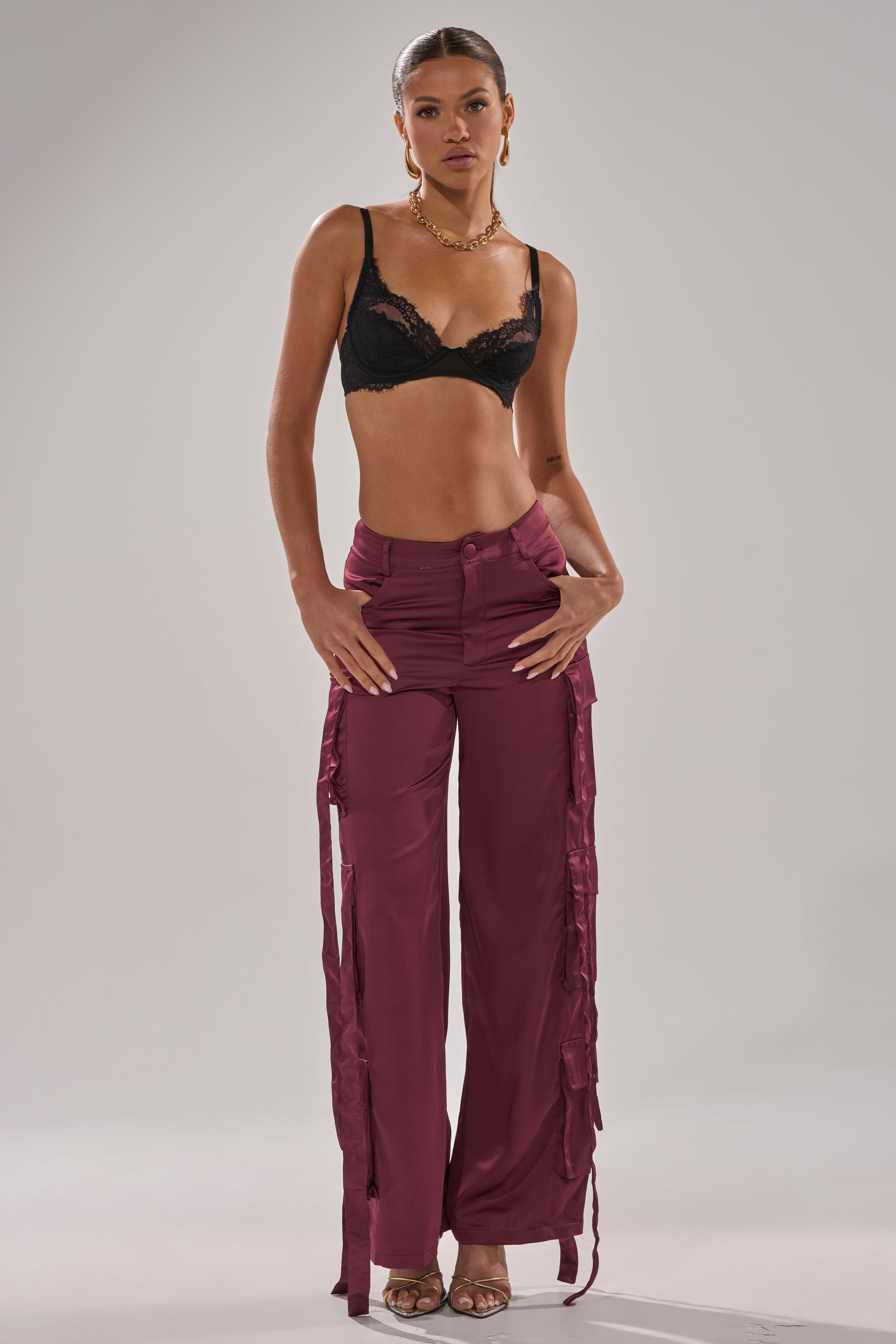 A woman poses against a plain background in the TAKE ME EVERYWHERE SATIN CARGO PANT, featuring a high-rise fit, wide-leg silhouette, and side ties, paired with a black lace bralette, gold hoop earrings, chunky necklace, and beige heeled sandals. 7
