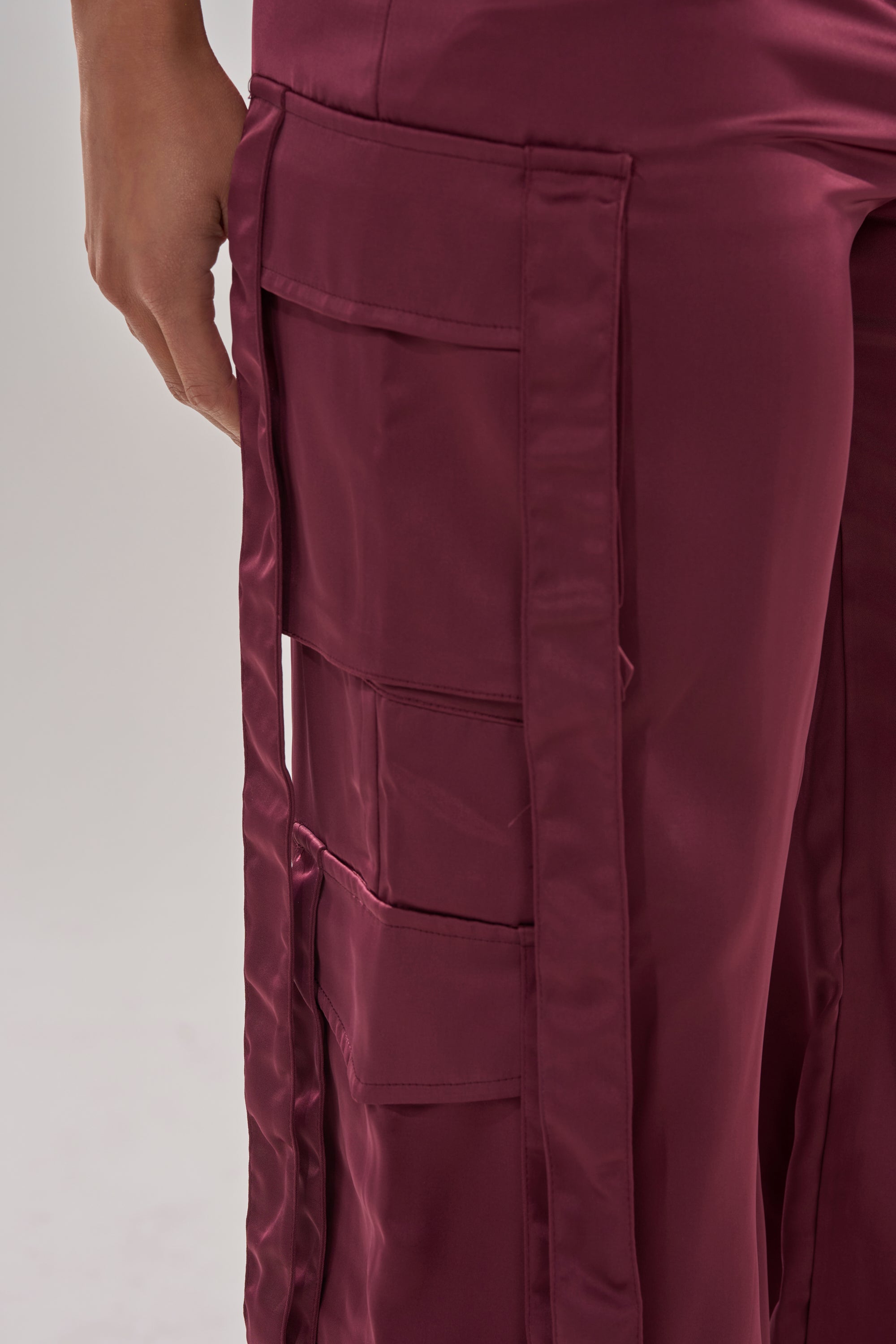 Close-up of a person wearing the TAKE ME EVERYWHERE SATIN CARGO PANT in shiny maroon with a high-rise waist, large side pockets, and stitched details. Their hand rests near the top pocket against a plain light background. 8
