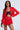 A woman in a bold red outfit with pleated skirt and the LILA CROPPED SATIN BLAZER featuring a notch lapel collar, revealing a black lace top. She wears star and moon pendant necklace, has long hair, and stands confidently against a light background.