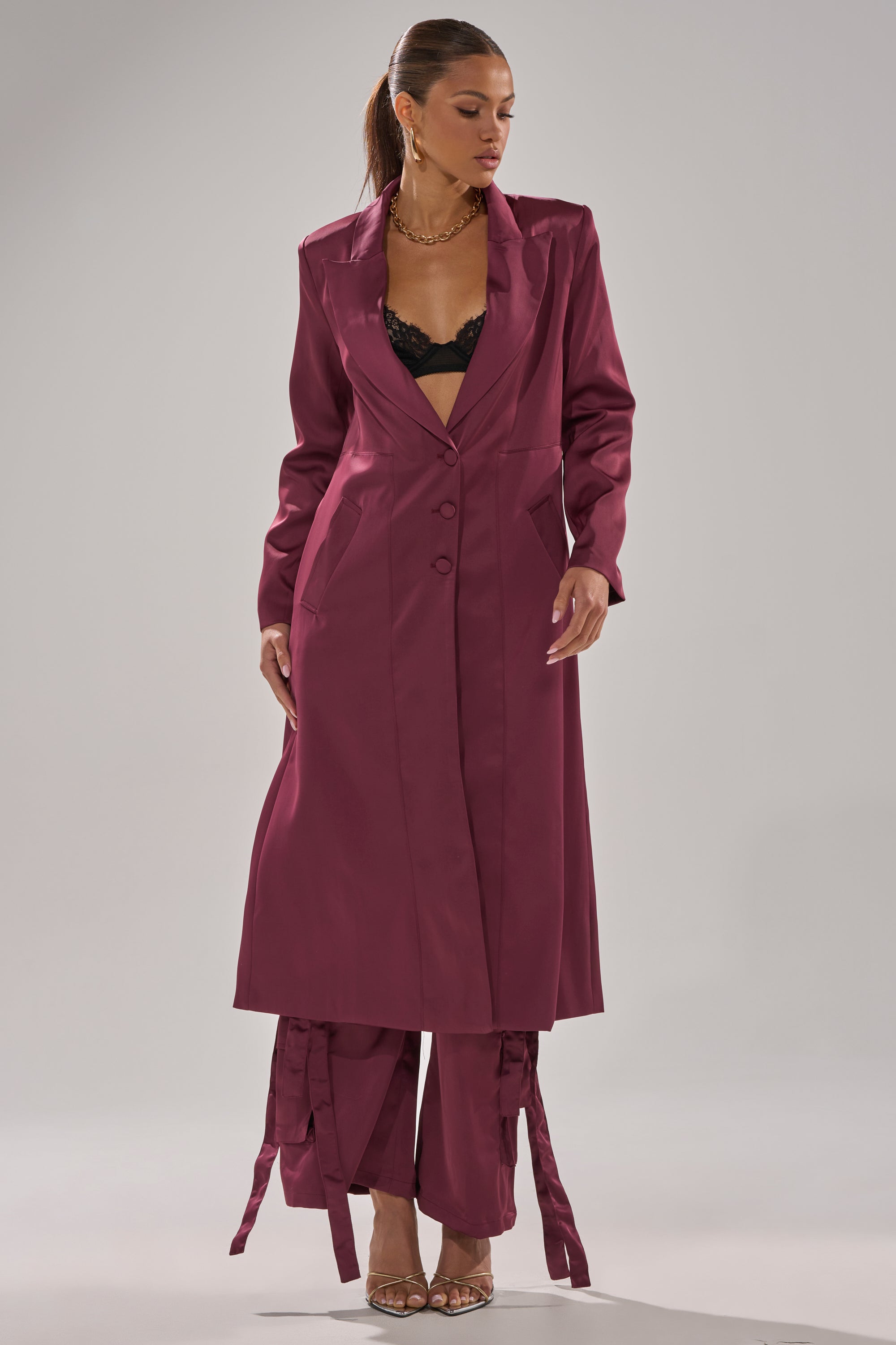 A woman styles the TAKE ME EVERYWHERE SATIN TRENCH IN BURGUNDY over matching trousers and a black lace bra, accessorized with gold jewelry and heeled sandals. Her hair is pulled back for a polished look. 0