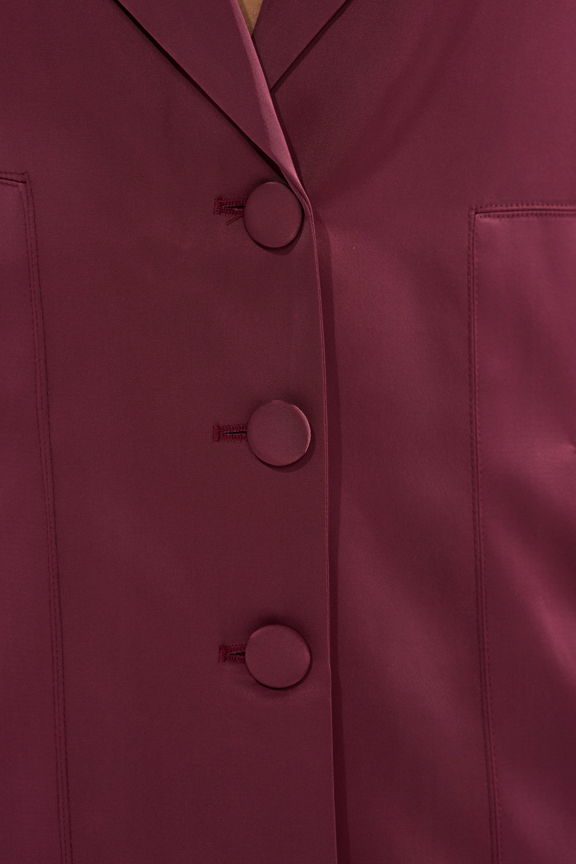 Close-up of the TAKE ME EVERYWHERE SATIN TRENCH IN BURGUNDY, showcasing three large fabric-covered buttons and two pocket seams flanking the buttons. 9