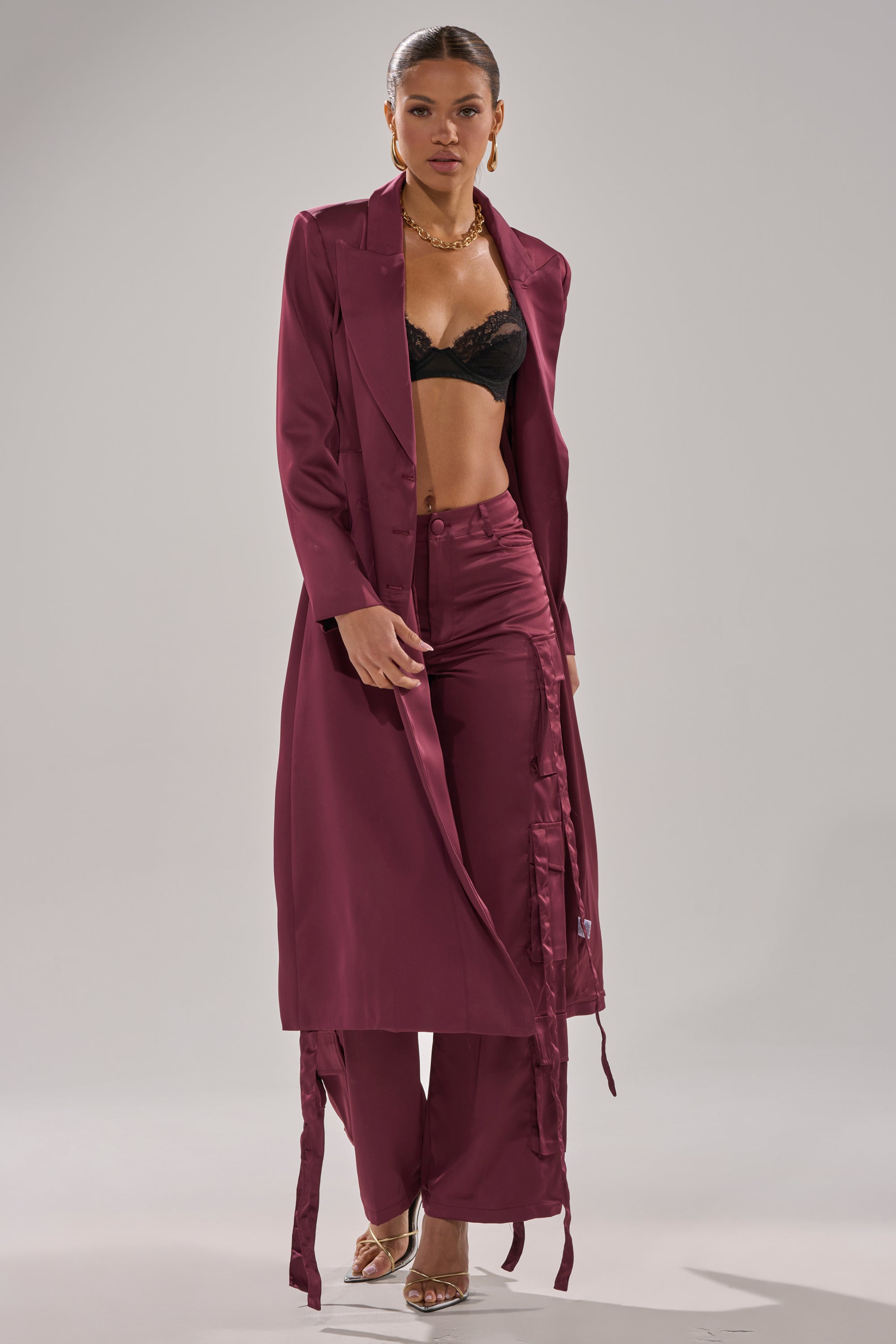 A woman models the TAKE ME EVERYWHERE SATIN TRENCH IN BURGUNDY with matching high-waisted pants, a black lace bralette, gold jewelry, and strappy heels against a plain light gray background. 1