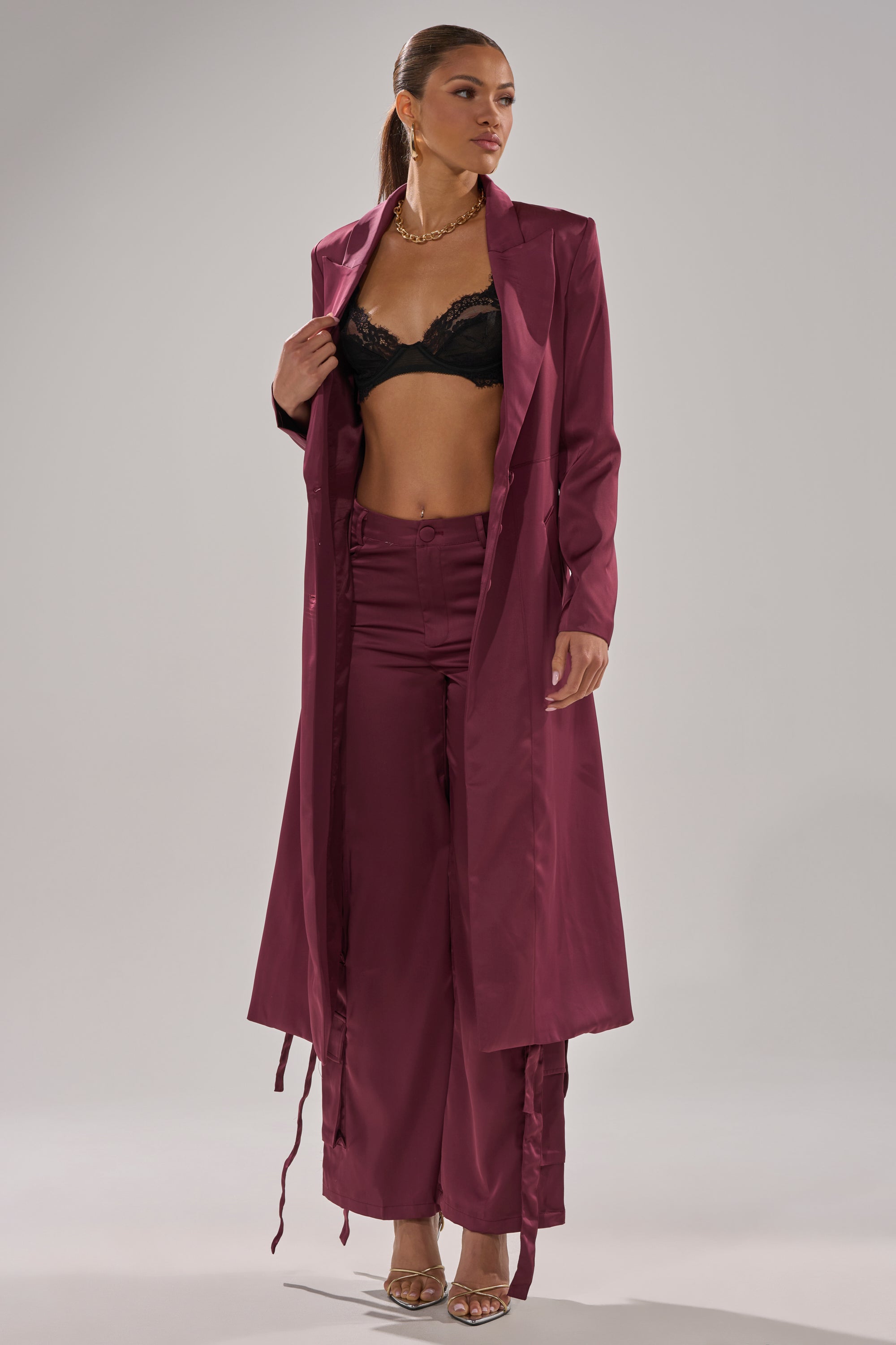 A woman poses in the TAKE ME EVERYWHERE SATIN TRENCH IN BURGUNDY, with matching wide-leg pants, a black lace bralette, gold necklace, and heeled sandals against a plain light gray background. 3