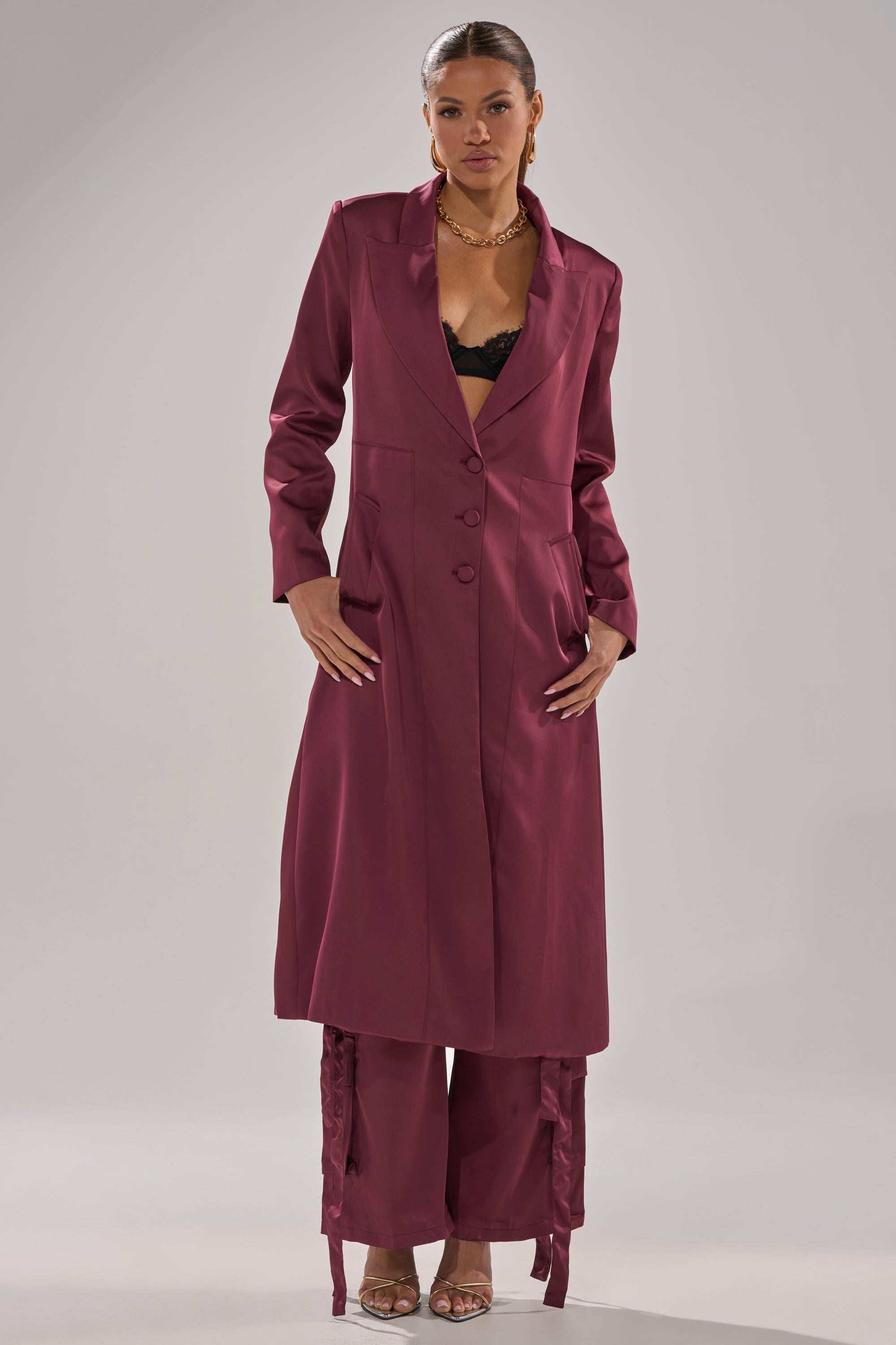 Wearing the TAKE ME EVERYWHERE SATIN TRENCH IN BURGUNDY over matching wide-leg pants and a black lace top, this look is styled with gold jewelry and open-toe heels against a plain light background. 4