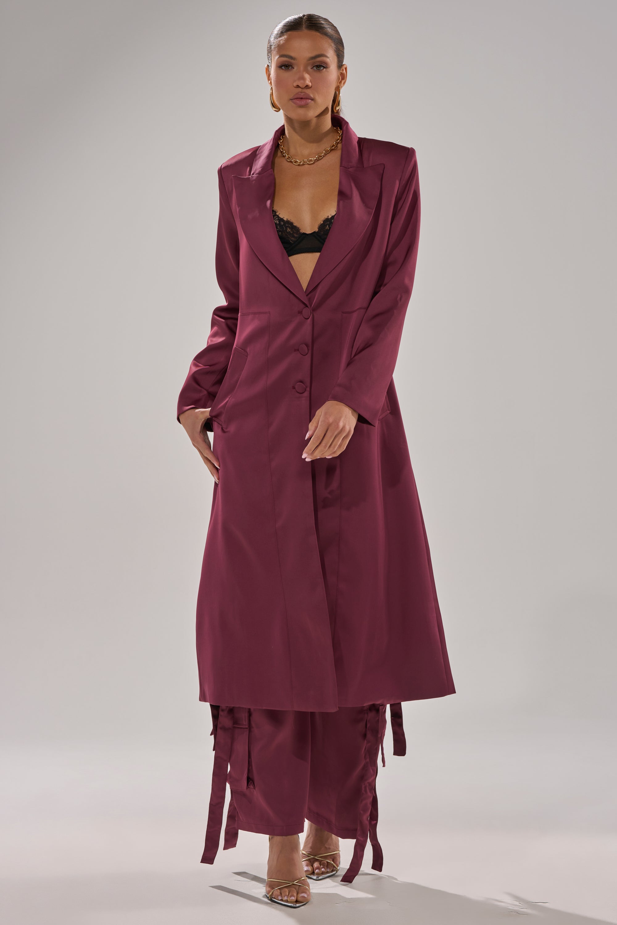 A woman poses confidently against a light gray background wearing the TAKE ME EVERYWHERE SATIN TRENCH IN BURGUNDY over a matching skirt and black lace bra, accessorized with gold jewelry and clear high heels. 5