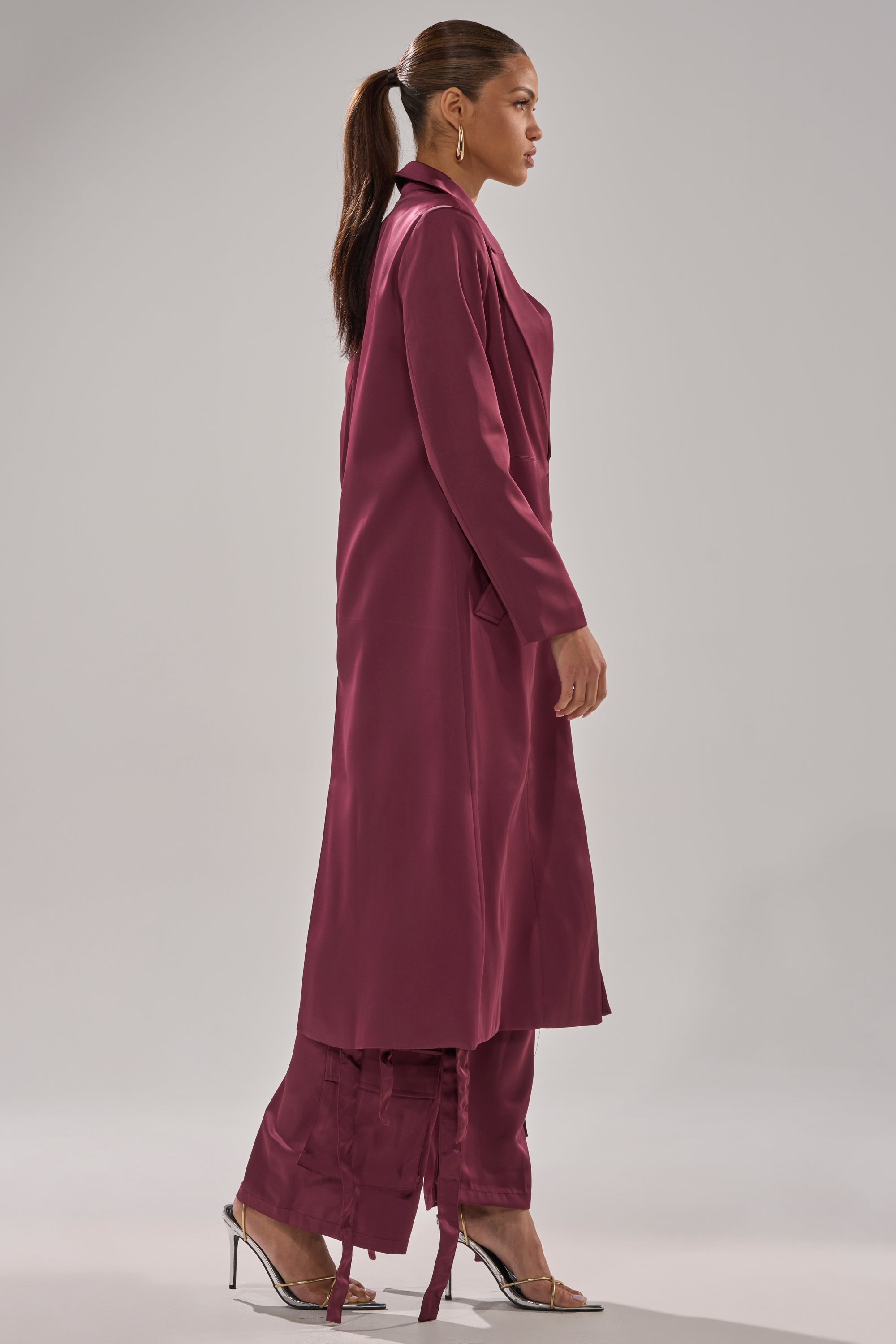 A woman in profile wears the TAKE ME EVERYWHERE SATIN TRENCH IN BURGUNDY with matching wide-legged pants featuring tied hems, clear high-heeled sandals, and gold hoop earrings. Her hair is styled in a ponytail. 6