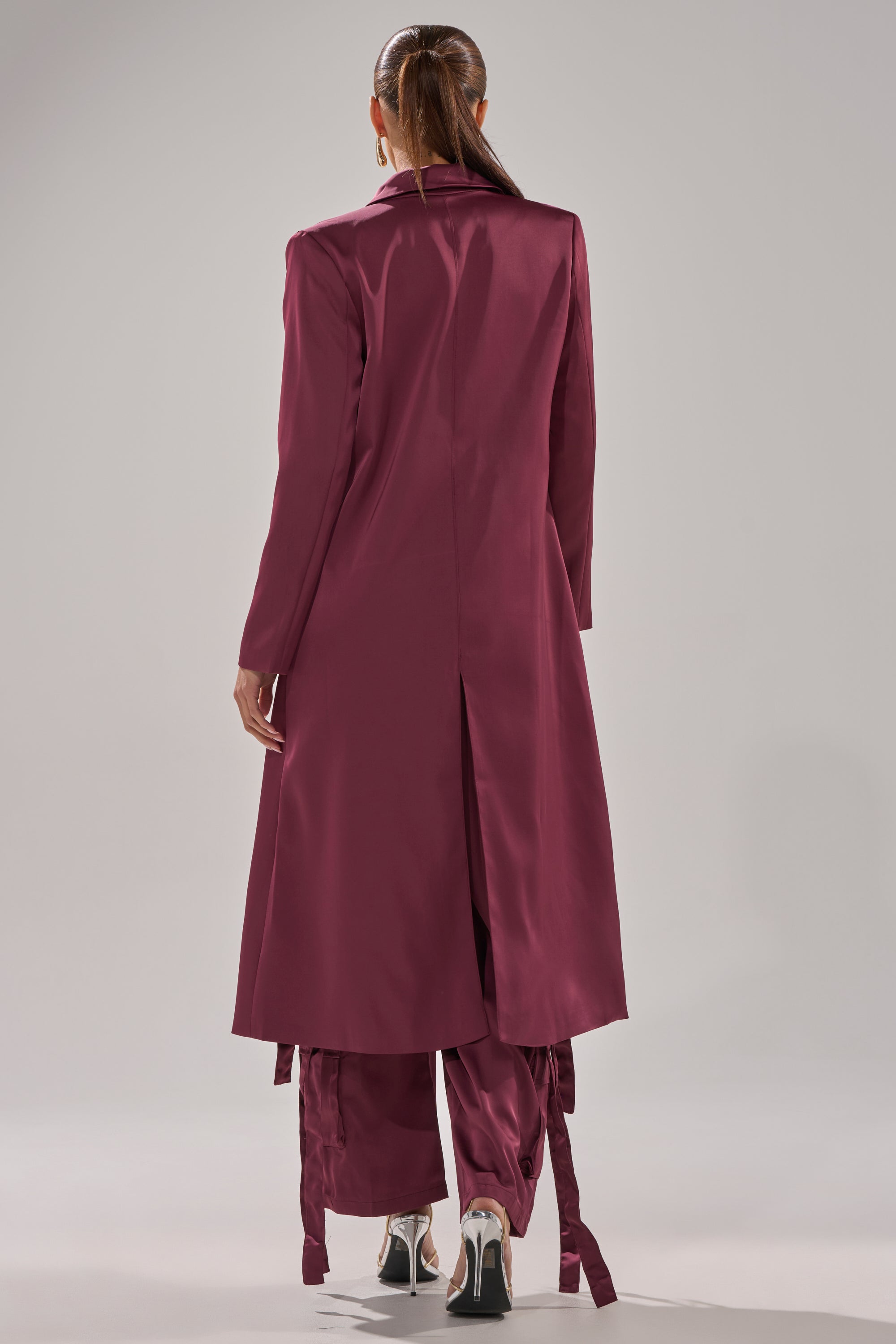 A woman with long brown hair in a ponytail models the TAKE ME EVERYWHERE SATIN TRENCH IN BURGUNDY with matching pants and shiny silver heels, seen from behind against a light gray background, showcasing the elegance of this look. 7