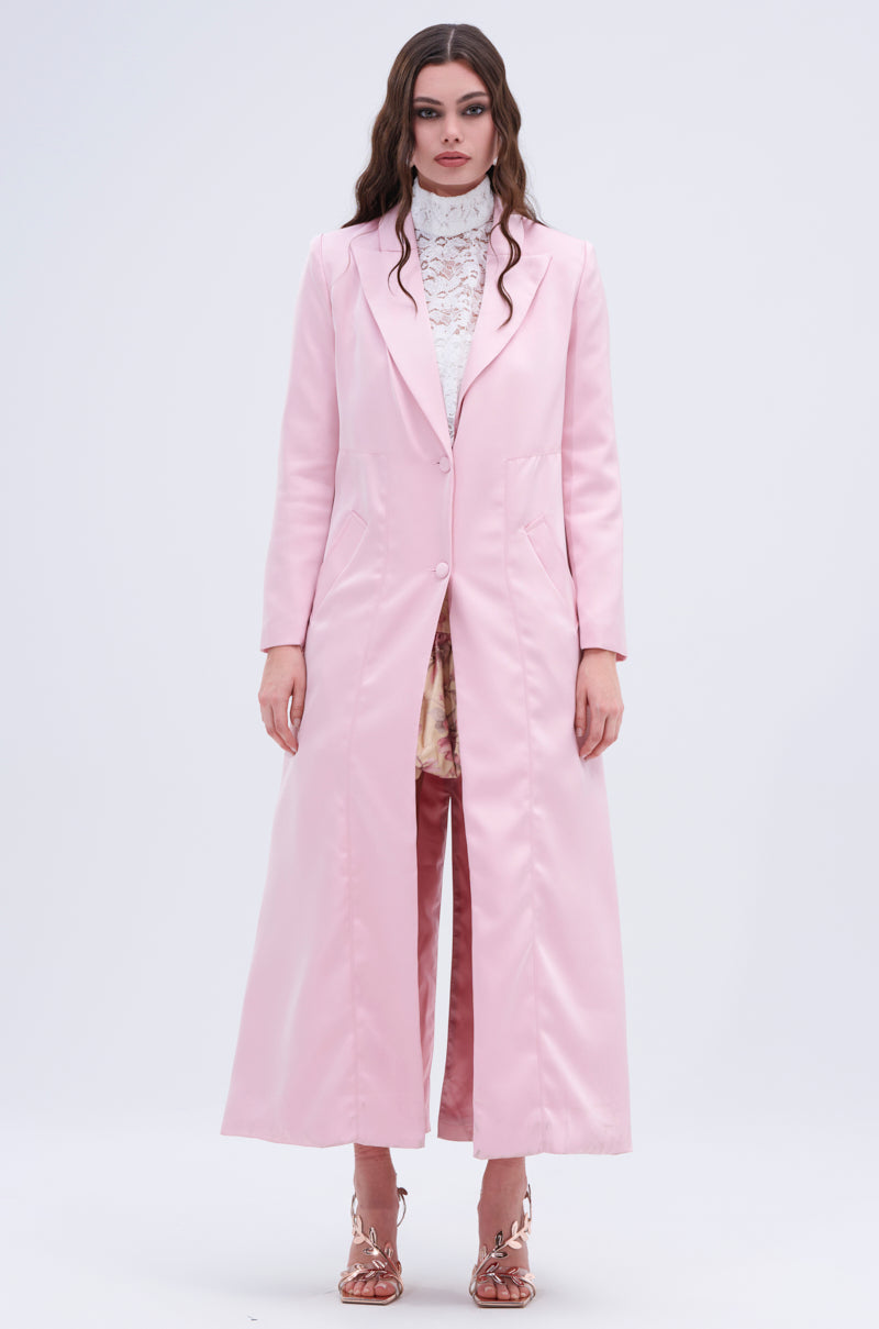 A woman stands against a plain background wearing the "TAKE ME EVERYWHERE SATIN TRENCH" over a white lace top, paired with pink trousers and strappy heels. Her hair is styled in loose waves, and she maintains a neutral expression.