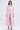 A woman stands against a plain background wearing the "TAKE ME EVERYWHERE SATIN TRENCH" over a white lace top, paired with pink trousers and strappy heels. Her hair is styled in loose waves, and she maintains a neutral expression.