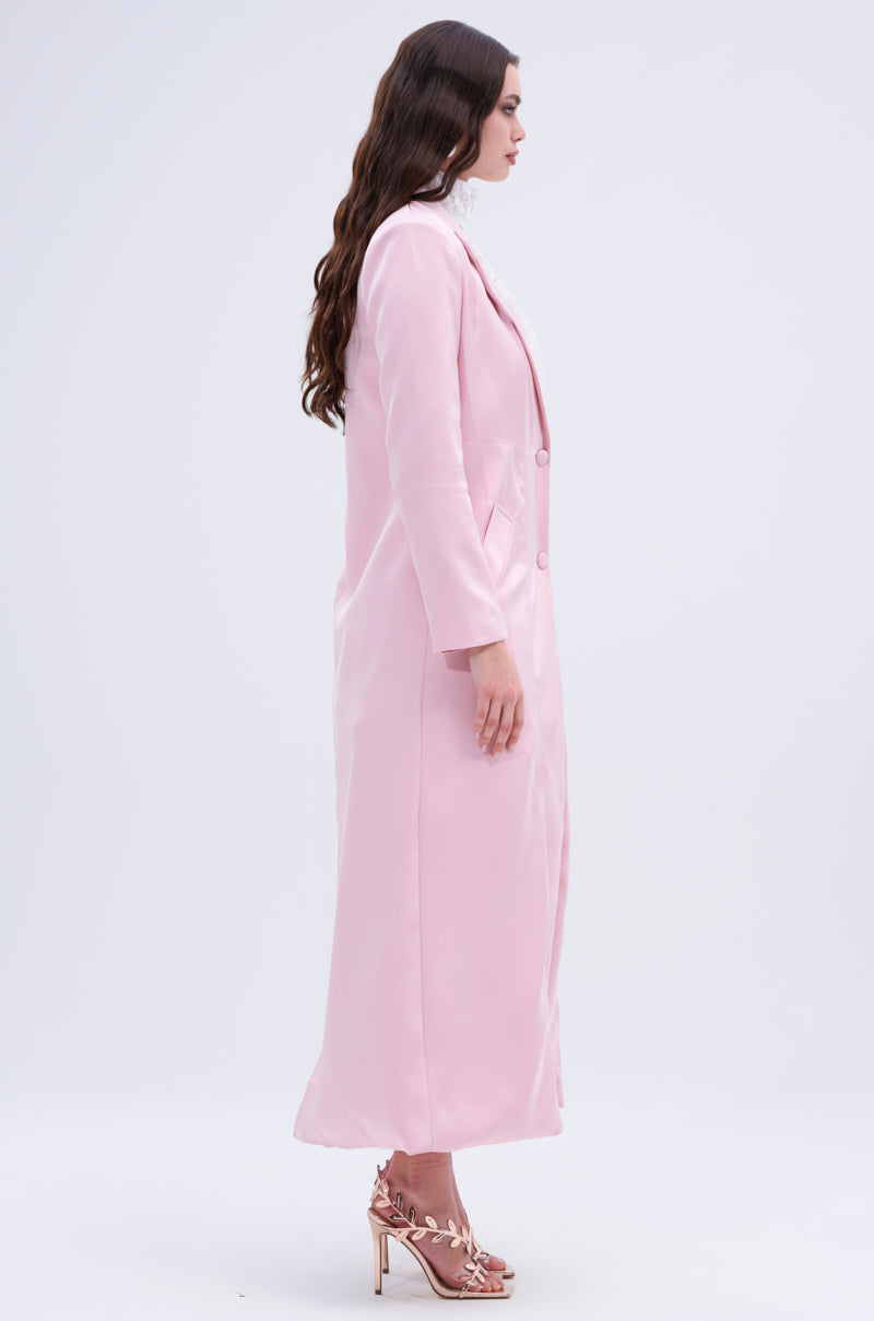 A woman in profile wears the luxurious TAKE ME EVERYWHERE SATIN TRENCH, elegantly paired with high-heeled sandals featuring floral details. Her long, wavy brown hair cascades against a plain, light background.