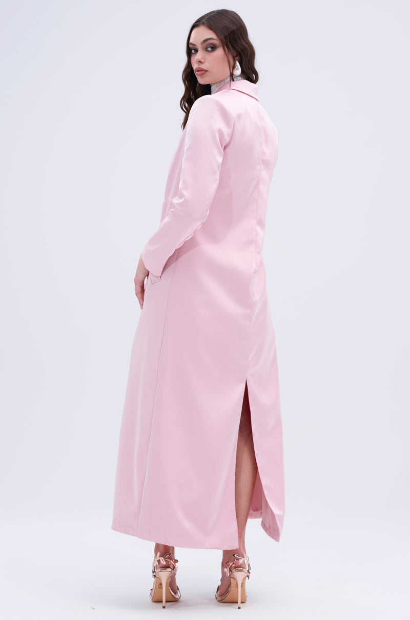 A woman with long hair stands facing slightly away, draped in the TAKE ME EVERYWHERE SATIN TRENCH from the AKIRA Label. The pink coat has a high back slit and pairs well with her high-heeled sandals, while the minimalist white background highlights her elegant style.