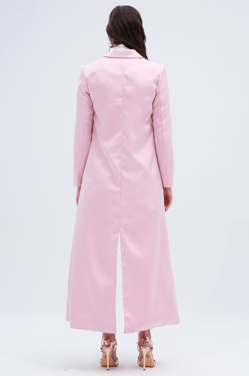 Wearing light-colored, strappy high heels, a person with long hair stands facing away in the TAKE ME EVERYWHERE SATIN TRENCH. This luxurious pastel pink coat features a back slit and reaches mid-calf against a plain light gray background.