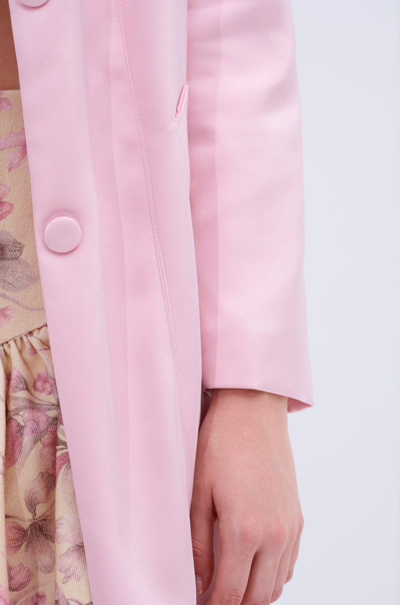 Close-up of a person wearing the light pink TAKE ME EVERYWHERE SATIN TRENCH with large buttons, partially covering a floral-patterned dress. The luxurious satin complements the ensemble as their hand rests at their side against a light gray backdrop.