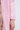Close-up of a person wearing the light pink TAKE ME EVERYWHERE SATIN TRENCH with large buttons, partially covering a floral-patterned dress. The luxurious satin complements the ensemble as their hand rests at their side against a light gray backdrop.