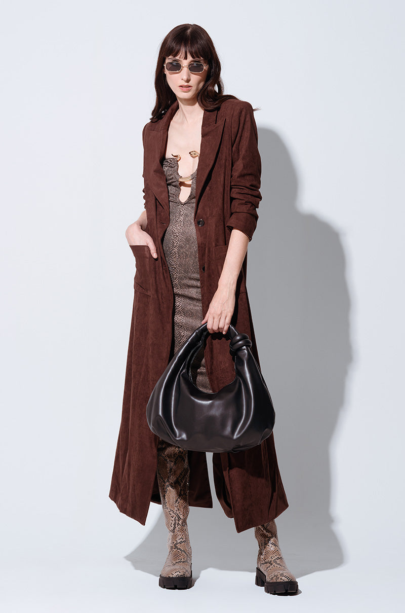 A person in sunglasses, a VERO SUEDE EFFECT TRENCH over a patterned dress, and knee-high snakeskin boots carries a large black bag. The maxi-length trench incorporates layered textures and earthy tones to enhance the ensemble.