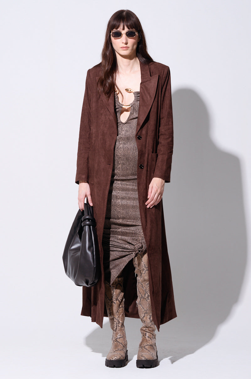 A person stands against a plain background wearing the VERO SUEDE EFFECT TRENCH in brown, a snake print dress, tall snakeskin boots, and sunglasses. They hold a black handbag.