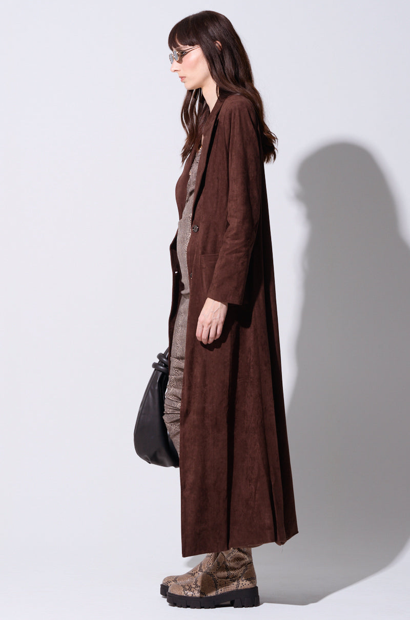 A person with long brown hair, wearing glasses and a VERO SUEDE EFFECT TRENCH over a gray outfit, holds a black bag. They stand in profile against a plain white background, highlighting their snakeskin-patterned platform boots.