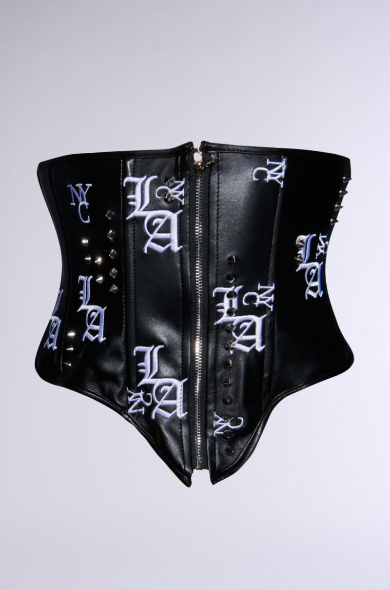 The "IN THE CITY EMBROIDERED UNDERBUST BELT" is a black faux leather corset-style top with white embroidered letters such as "LA" and "NYC," enhanced by metal studs and a central zipper on a structured, fitted design against a plain white background.