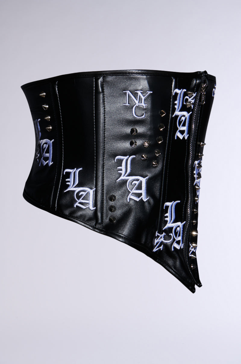 The IN THE CITY EMBROIDERED UNDERBUST BELT features a black faux leather corset-inspired design with white "LA" embroidery and metal studs. Its asymmetrical shape, glossy finish, and side lacing create a unique style against a plain white background.