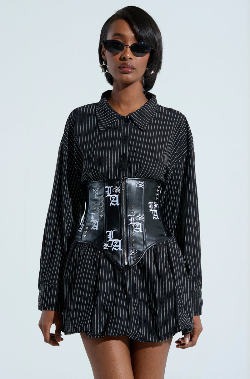 A person is wearing a black pinstripe dress with the "IN THE CITY EMBROIDERED UNDERBUST BELT" featuring white lettering. They have short hair, dark sunglasses, and stand against a light neutral background, adding contrast to their chic ensemble.