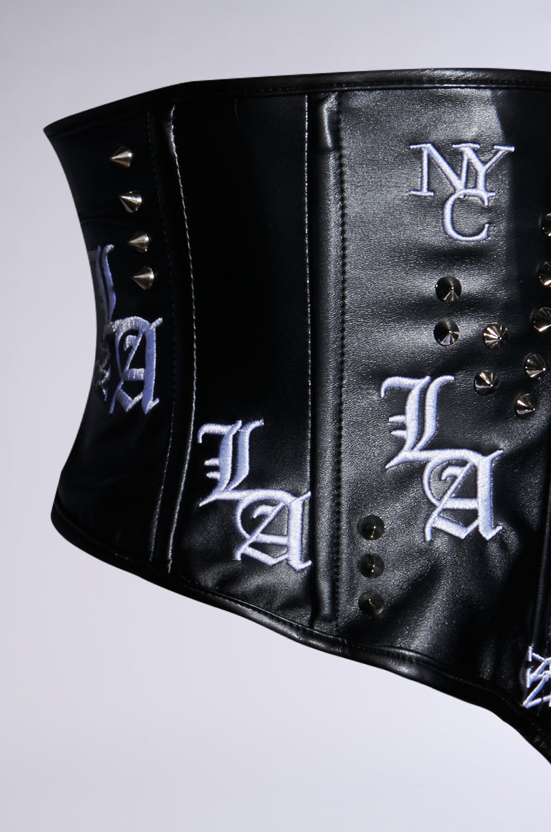 The IN THE CITY EMBROIDERED UNDERBUST BELT is a black faux leather corset-inspired piece with silver studs and white embroidered "NYC" and "LA" on the sides, combining urban fashion elements for an edgy style.