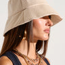 A woman with long brown hair wears the CHAMPAGNE SIPPIN BLING BUCKET HAT adorned with tonal rhinestones, large hoop earrings, and layered gold chain necklaces. She is dressed in a dark sleeveless top, against a plain light-colored background.