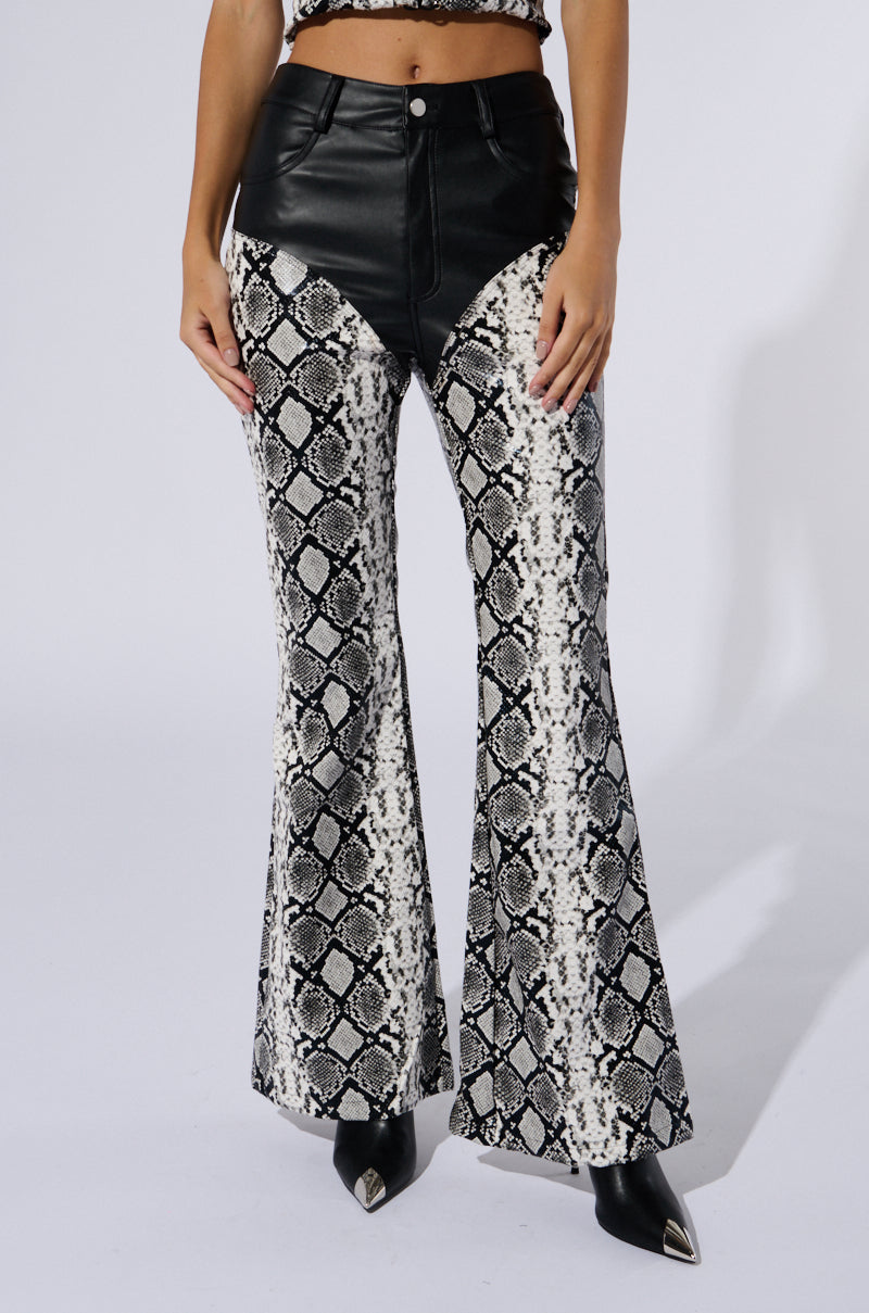 A person is wearing the Day and Night Trouser, a pair of high-waisted, black faux leather bell-bottom pants featuring a bold faux snakeskin print on the legs. They are paired with pointed black shoes. The background is plain white, and the person's upper body is not fully visible.