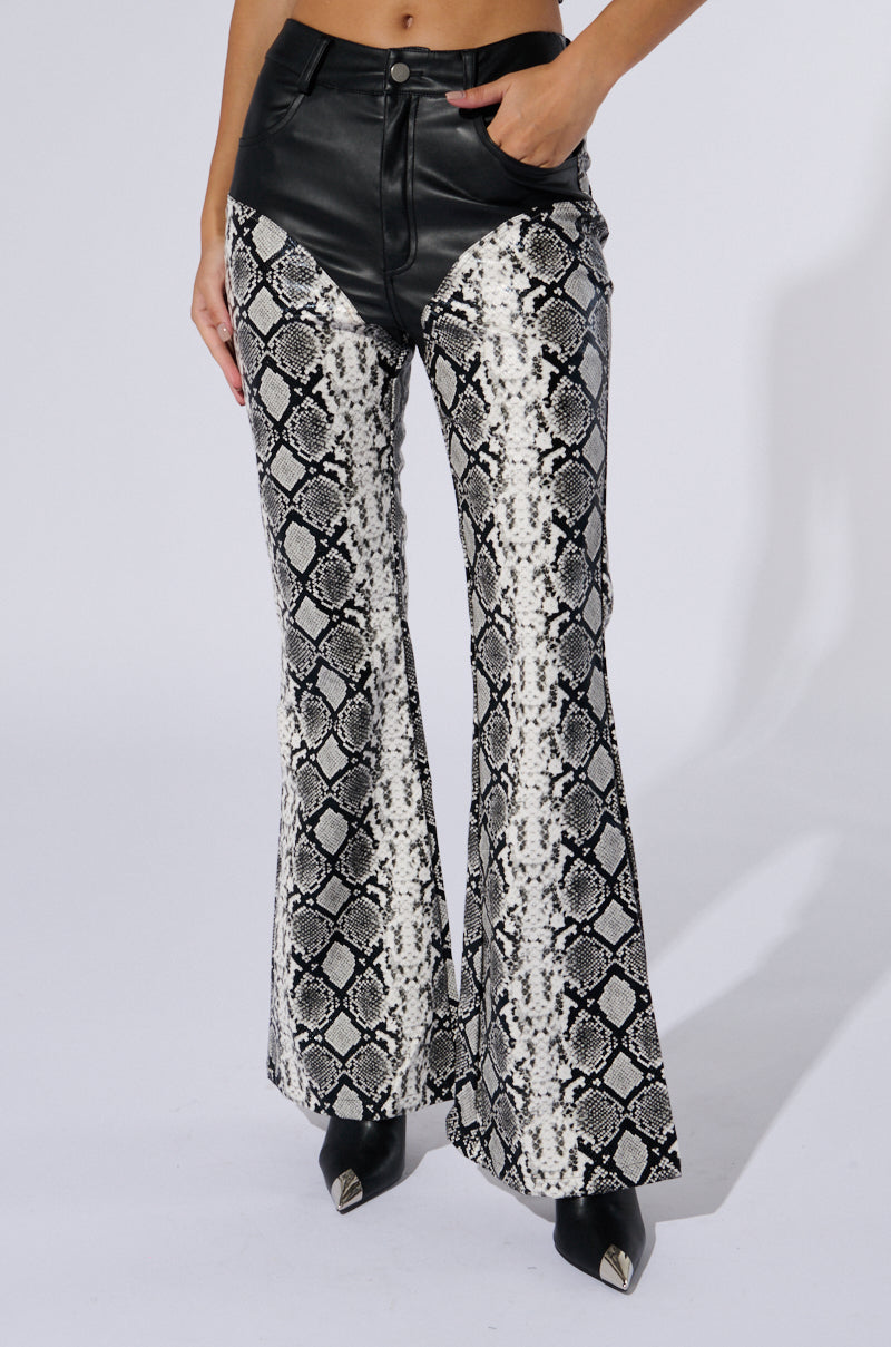 A person is dressed in the DAY AND NIGHT TROUSER, featuring black and white faux snakeskin flared pants with chap-inspired leather paneling around the waist. The high-waisted trousers are paired with black pointed shoes, and the person’s hands rest casually in their pockets against a plain white background.