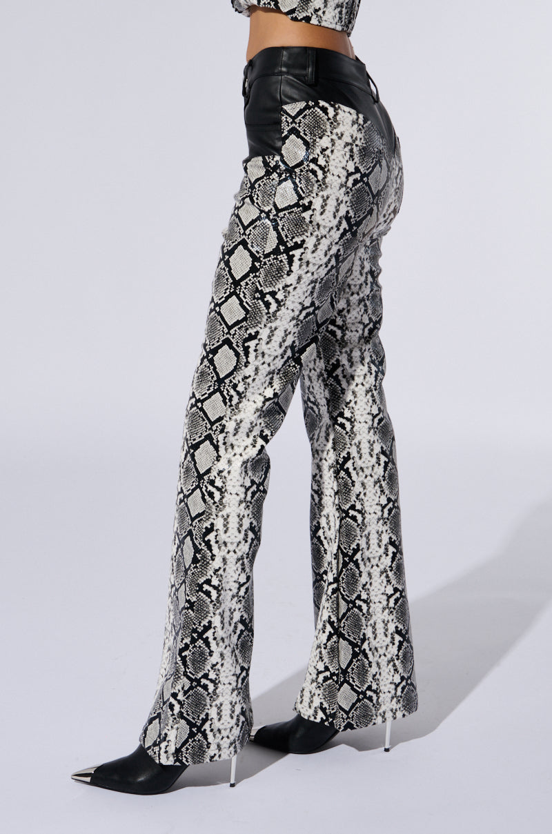 A person is modeling the "DAY AND NIGHT TROUSER," which features a high-waisted cut in a striking black and white faux snakeskin design. The trousers boast a fitted waist and hips, complemented by flared legs with stylish chap-inspired paneling. They are paired with black high-heeled pointy shoes against a plain white background.