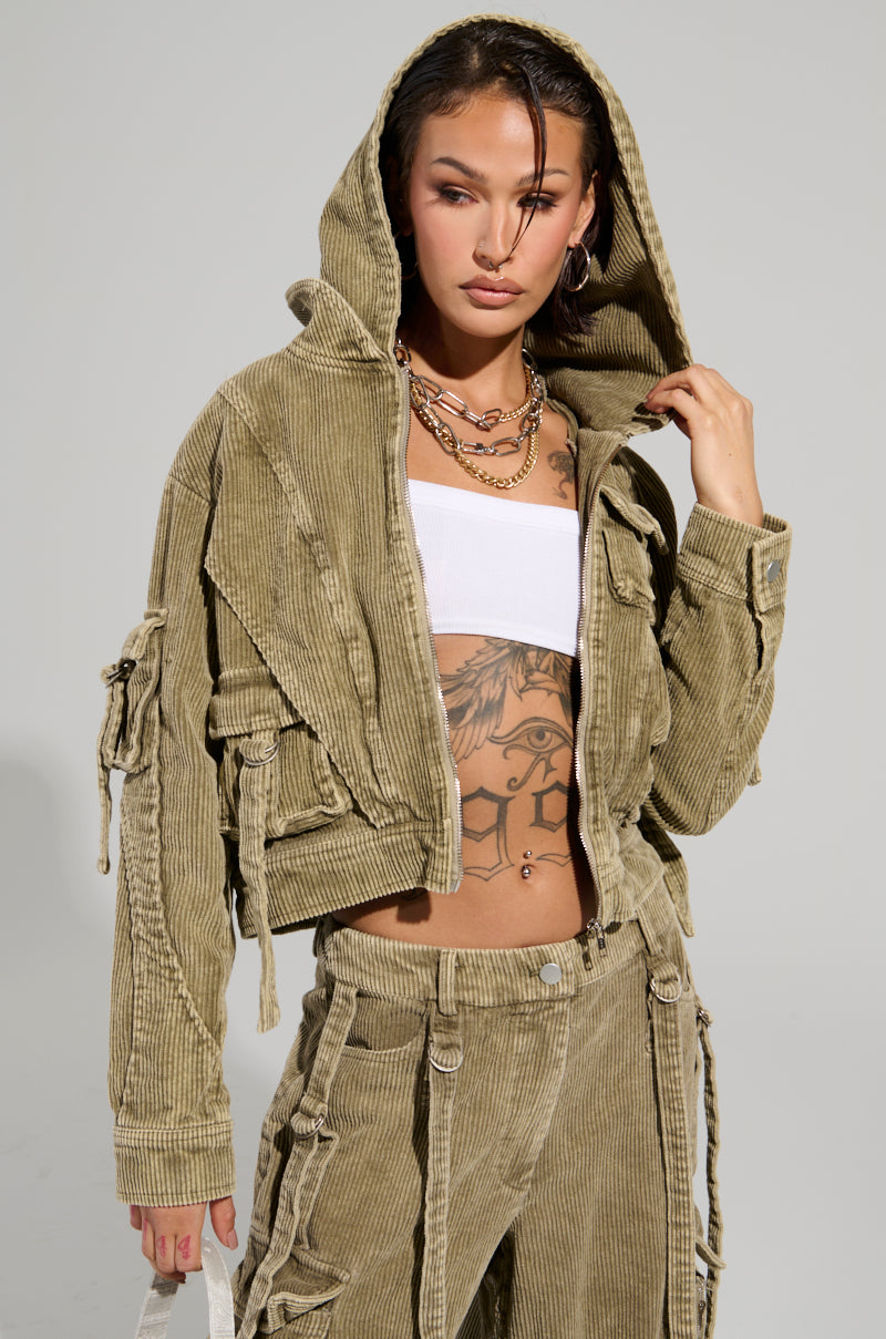 A person with tattoos is dressed in an AKIRA Label Stealthy Corduroy Cargo Bomber jacket and matching Corduroy Cargo Pants. They have short hair, hoop earrings, and layered necklaces. The jacket is slightly open, revealing a white top underneath.