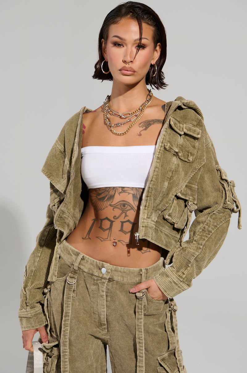 A person with shoulder-length hair poses confidently in an AKIRA Label ensemble, featuring the STEALTHY CORDUROY CARGO BOMBER. They wear a green corduroy bomber jacket and pants, with a white crop top underneath. Tattoos adorn their torso as they accessorize with hoop earrings and a layered chain necklace.