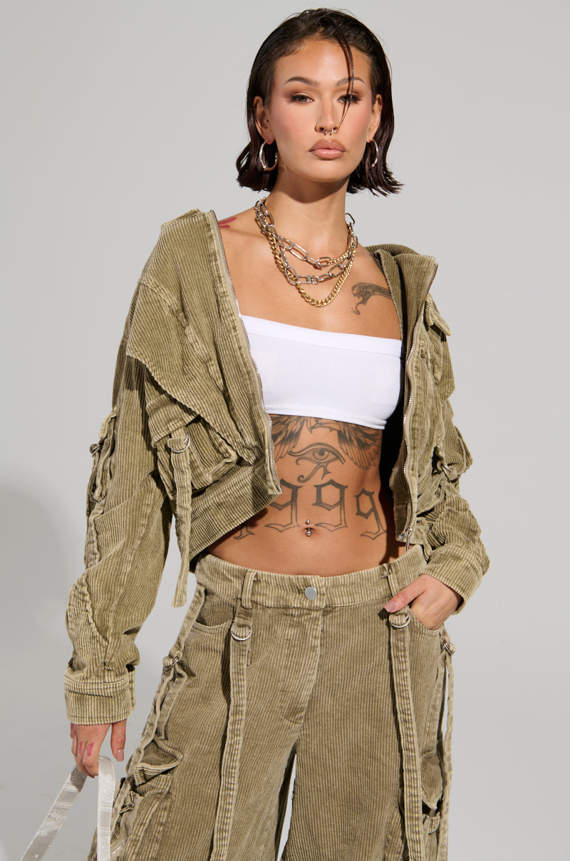 A person with short dark hair is rocking the AKIRA Label STEALTHY CORDUROY CARGO BOMBER, featuring a light olive-green cropped jacket combined with corduroy cargo pants. Tattoos adorn their chest and stomach, while gold hoop earrings and layered necklaces add a stylish edge.