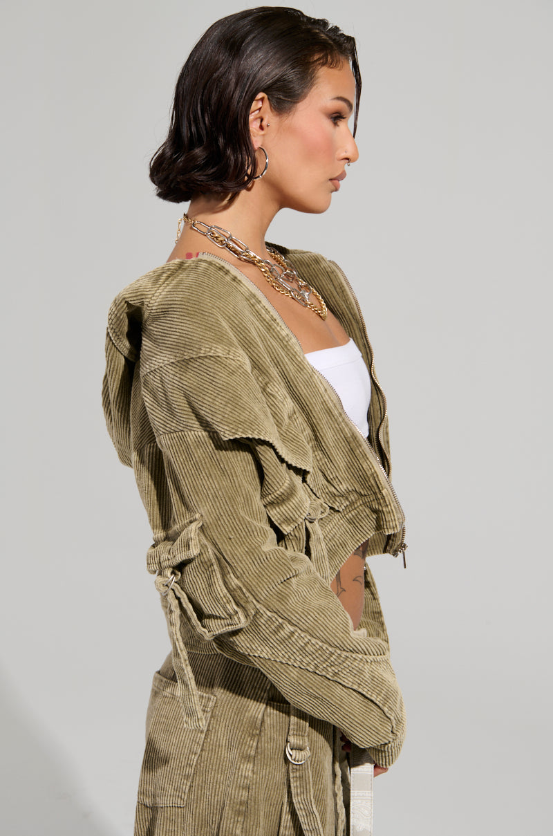 A person with short dark hair is wearing a light green AKIRA Label Stealthy Corduroy Cargo Bomber jacket, a white top, and layered necklaces. They are standing in profile against a plain background.