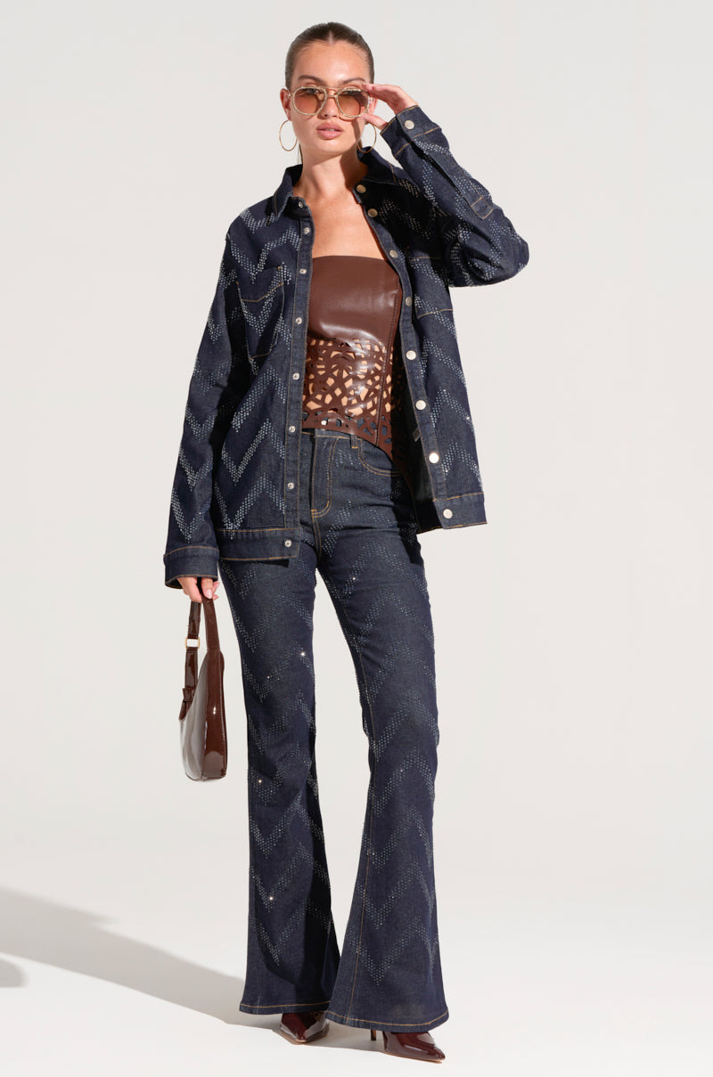 A person wearing sunglasses styles a dark wash denim jacket with zigzag patterns and GOOD KARMA EMBELLISHED FLARE JEANS. Underneath, they have on a brown patterned top, while carrying a small brown handbag and sporting brown pointed shoes.