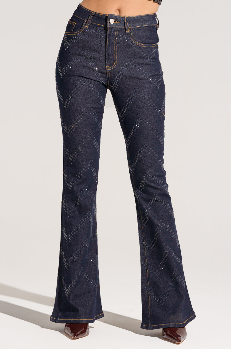 Someone is wearing the Good Karma Embellished Flare Jeans in a dark wash with a flared design and subtle patterned detailing. These jeans feature contrast stitching and a button closure, beautifully complemented by heeled shoes.