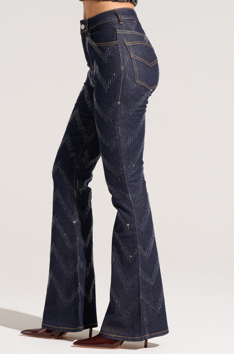 A person is wearing the GOOD KARMA EMBELLISHED FLARE JEANS in dark wash denim with a chevron pattern, paired with brown pointed-toe shoes on a plain, light background.