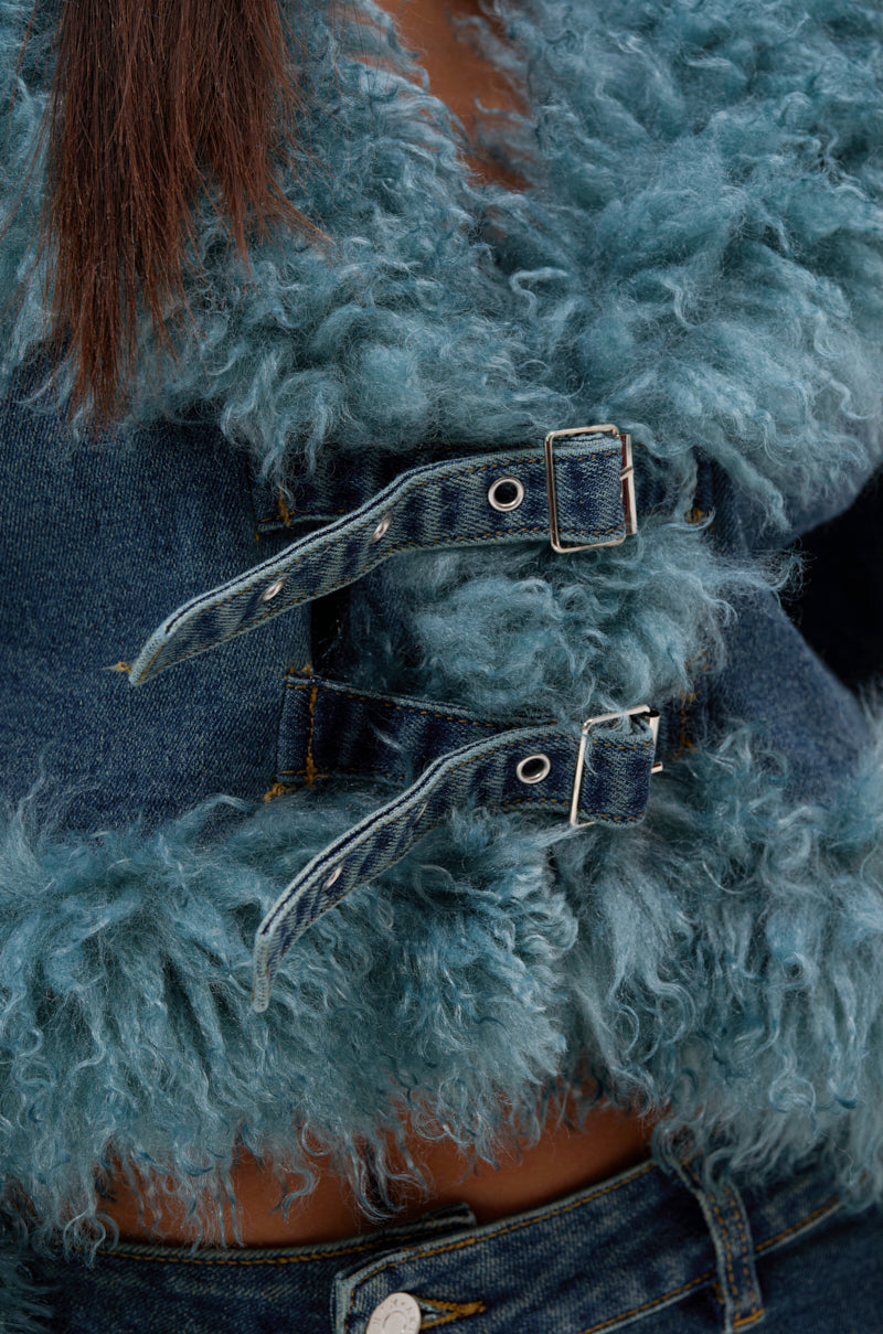 Close-up of the SIT PRETTY FUR TRIM DENIM JACKET, featuring blue denim with textured teal fur trim and two buckle details. The jacket’s hem and blue jeans waistband are visible, with long brown hair draped on the left.