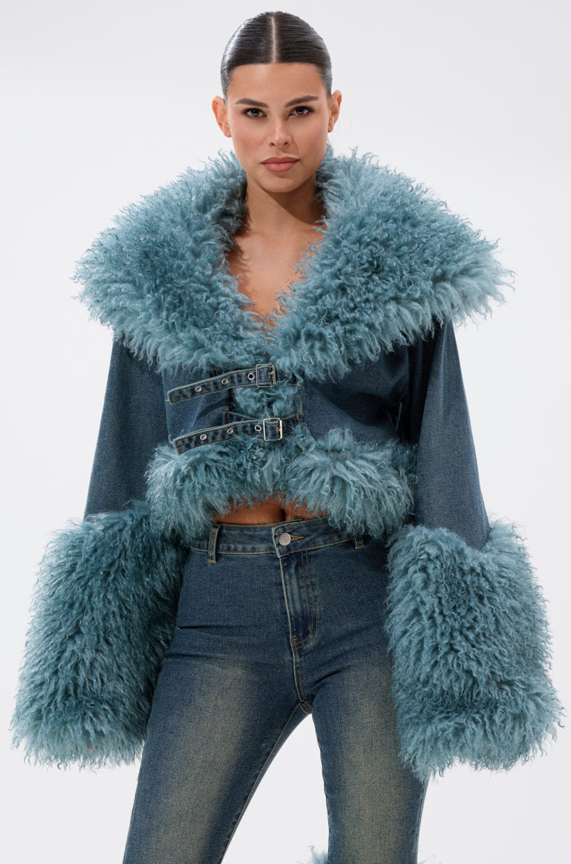 A woman wears the SIT PRETTY FUR TRIM DENIM JACKET, a cropped Y2K-style denim jacket with bold teal faux fur on the collar, cuffs, and waistband, plus two front buckle straps, paired with matching jeans against a plain background.