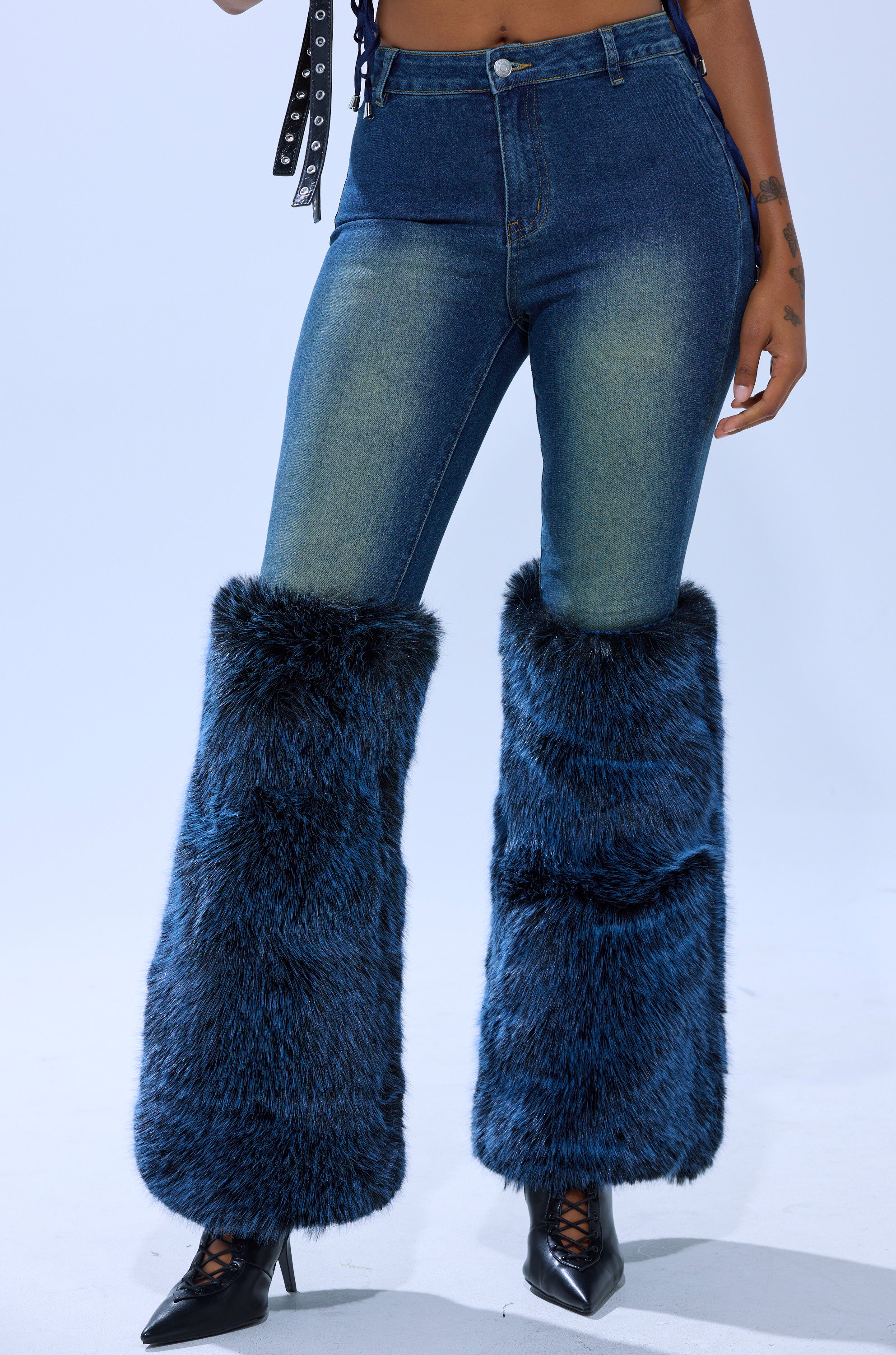 A person wears the ALL A DREAM DENIM PANT WITH FUR CUFFS—high-rise denim pants with bold blue faux fur cuffs on the lower legs—paired with black pointed-toe ankle boots. The background is plain light blue.